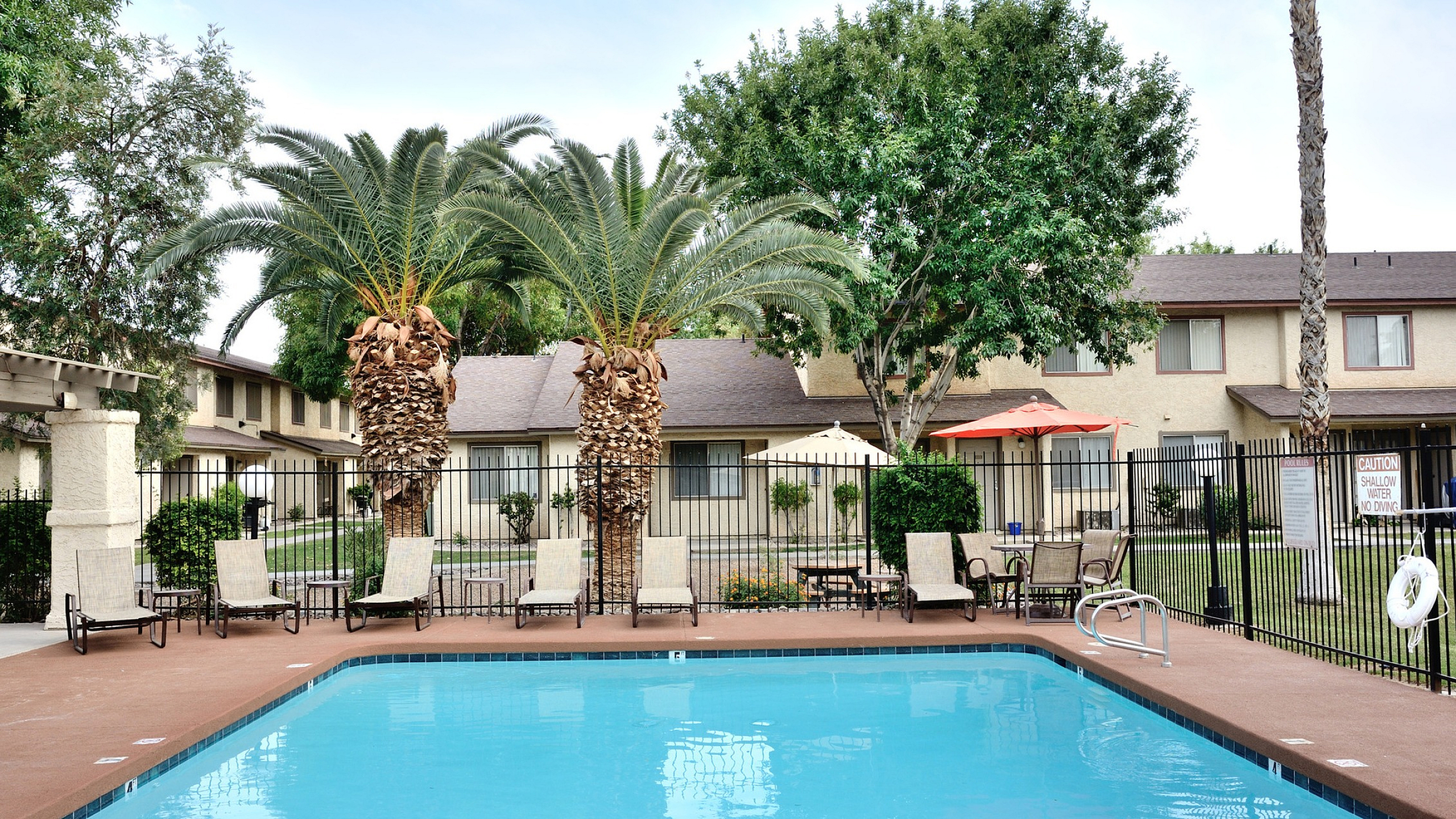 Inviting outdoor pool area at Camelot Apartments in Yuma, Arizona surrounded by lush greenery and palm trees, complemented by comfortable lounge chairs, perfect for relaxation.