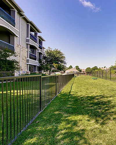 Charming apartment exterior with lush green dog park area, featuring playful obstacles and safe metal fencing under a clear blue sky.