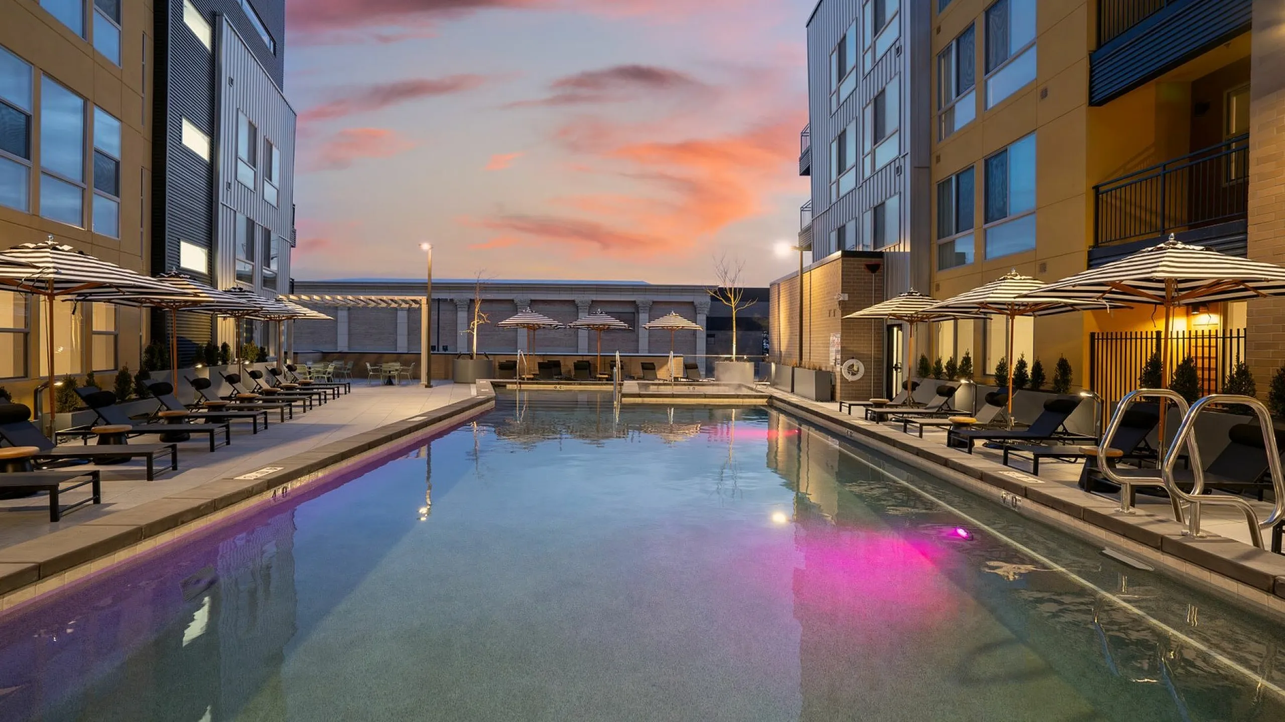 Luxury apartment complex showcasing a modern outdoor pool area, surrounded by sleek lounge chairs and stylish umbrellas, set against a stunning sunset backdrop.