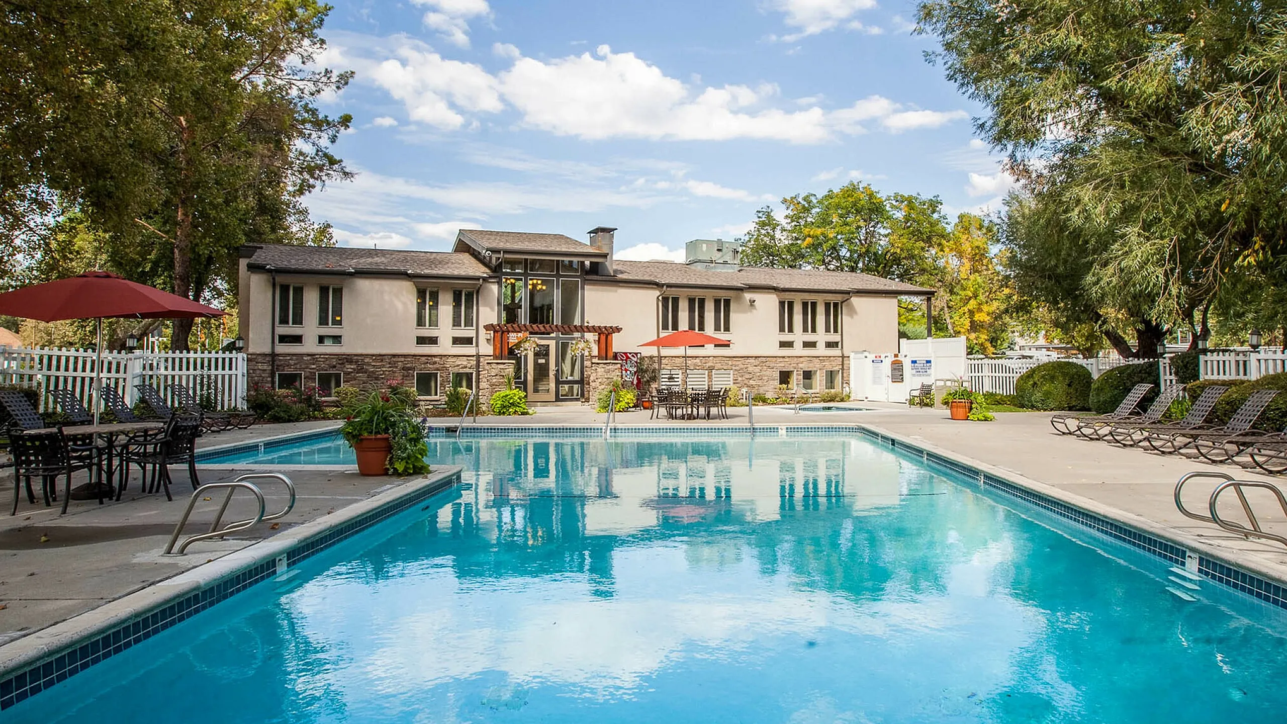 Inviting swimming pool area at Fox Point in Old Farm with lounge chairs and dining tables, surrounded by lush greenery and a charming residential building under a clear blue sky.