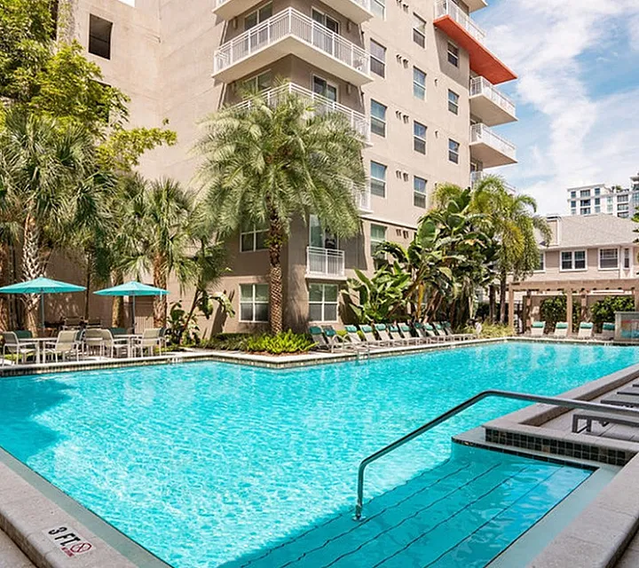 Luxurious swimming pool surrounded by tropical palm trees and modern apartment complex, featuring comfortable lounge chairs and shaded seating areas in Downtown St Pete