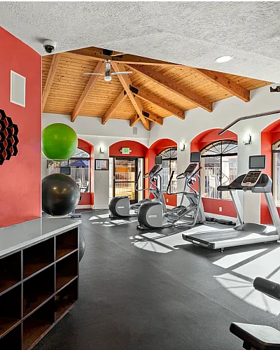 Bright, modern fitness center with state-of-the-art cardio equipment, vibrant red accents, and natural wood ceiling at Pavilions Apartments in Albuquerque
