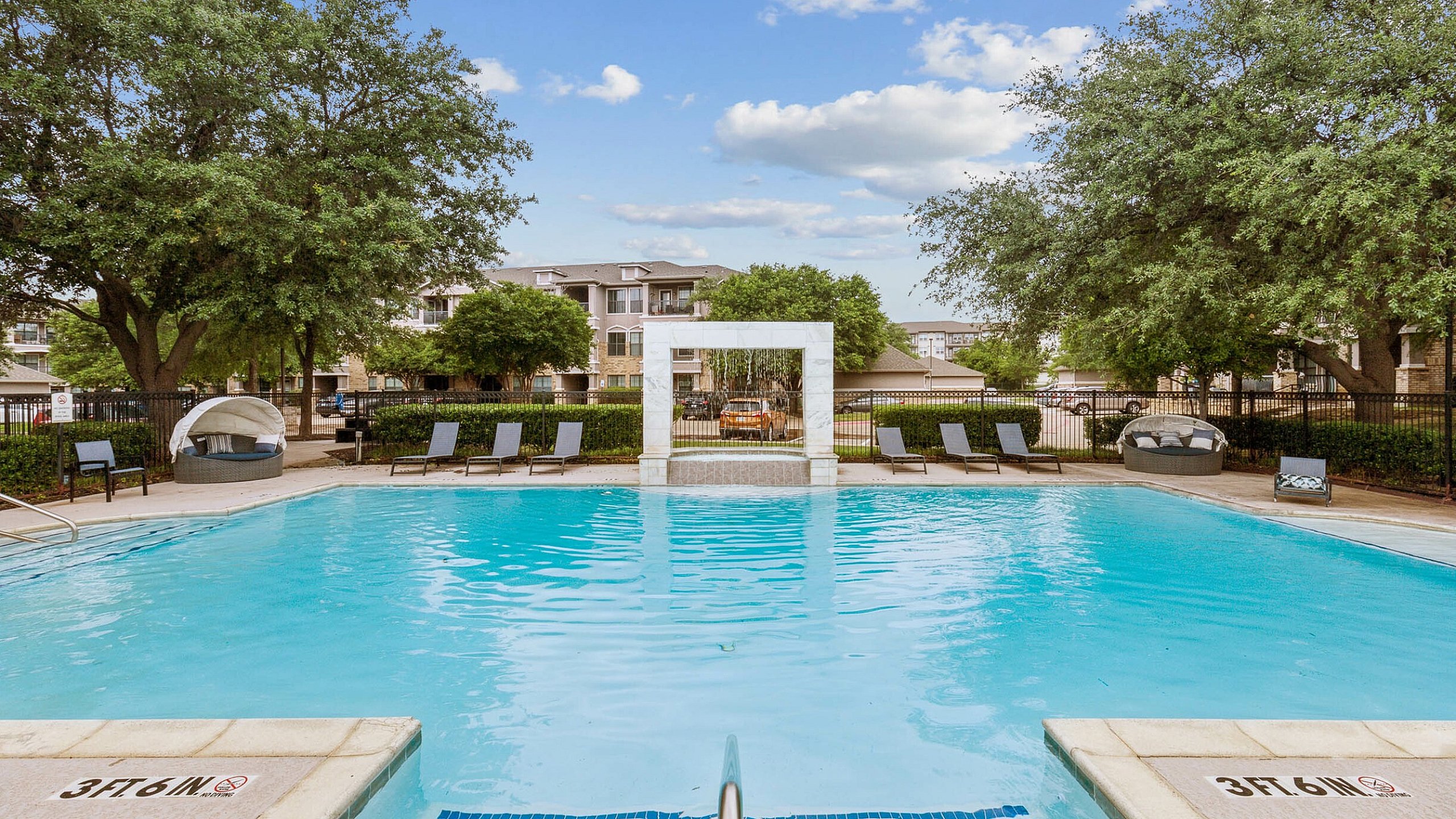 Luxurious apartment complex with a sparkling pool surrounded by lush greenery and stylish lounging areas.