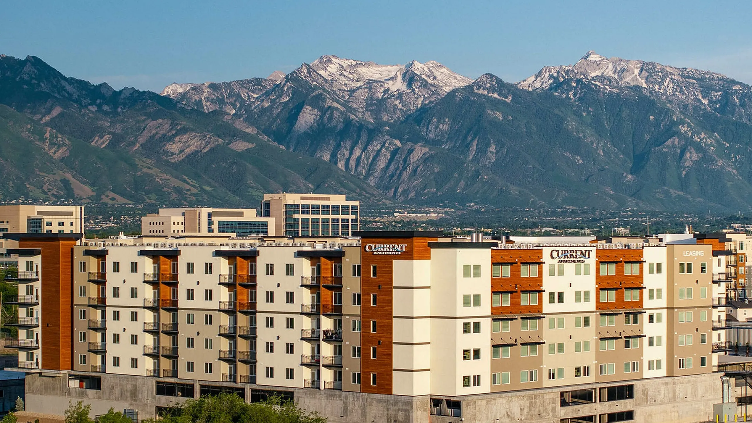 Modern apartment complex, Current Apartments, with stunning Wasatch Mountain views, featuring contemporary architecture and lush greenery below in Murray Utah