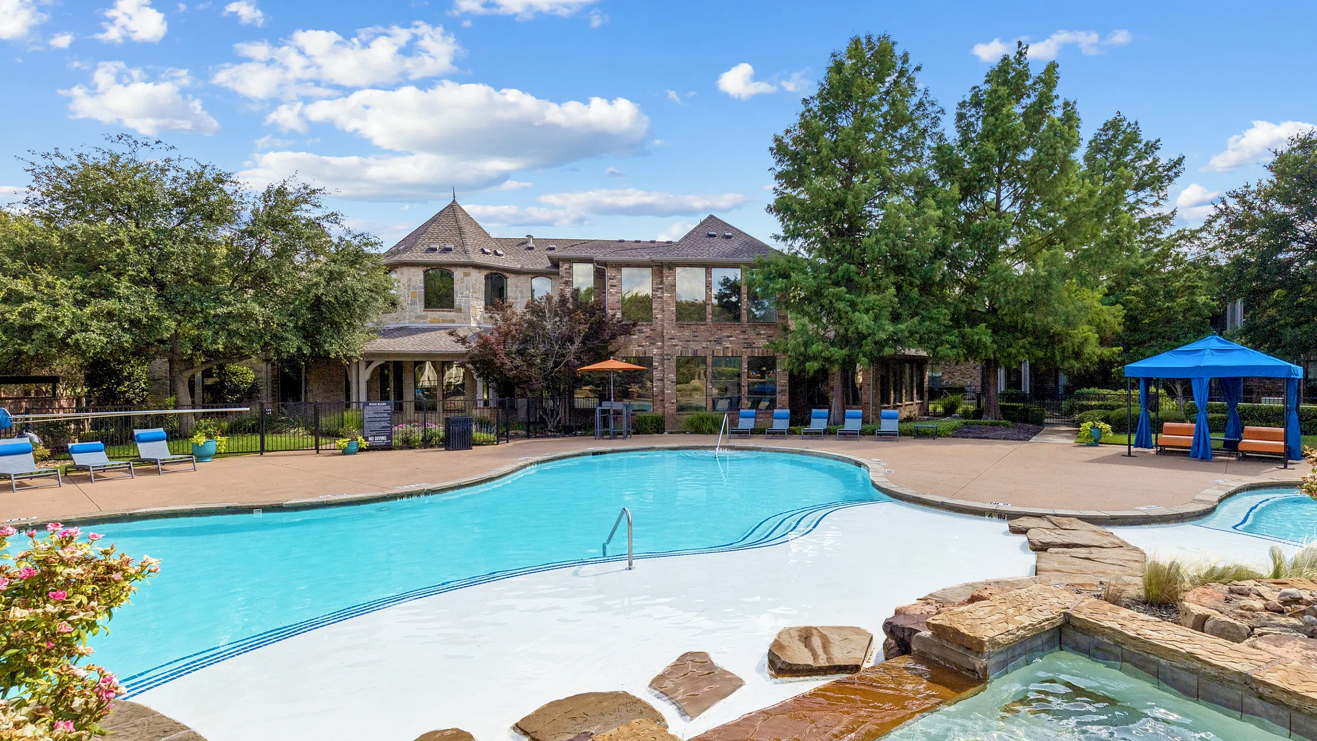 Luxurious apartment complex with a stunning resort-style pool, stylish cabanas, and lush garden settings against a clear blue sky backdrop.