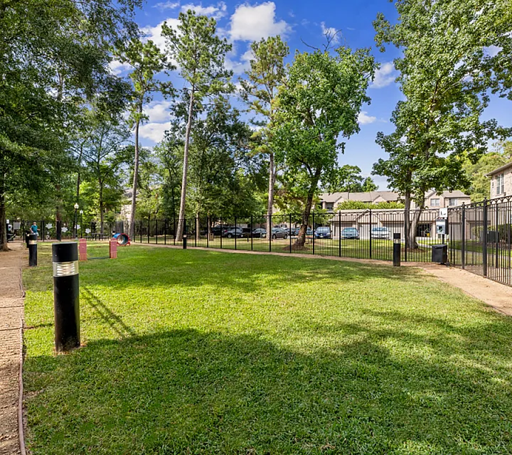 Charming community garden with lush green lawn, tree-lined paths, and cozy seating areas under a clear blue sky, perfect for relaxation Raveneaux Apartments