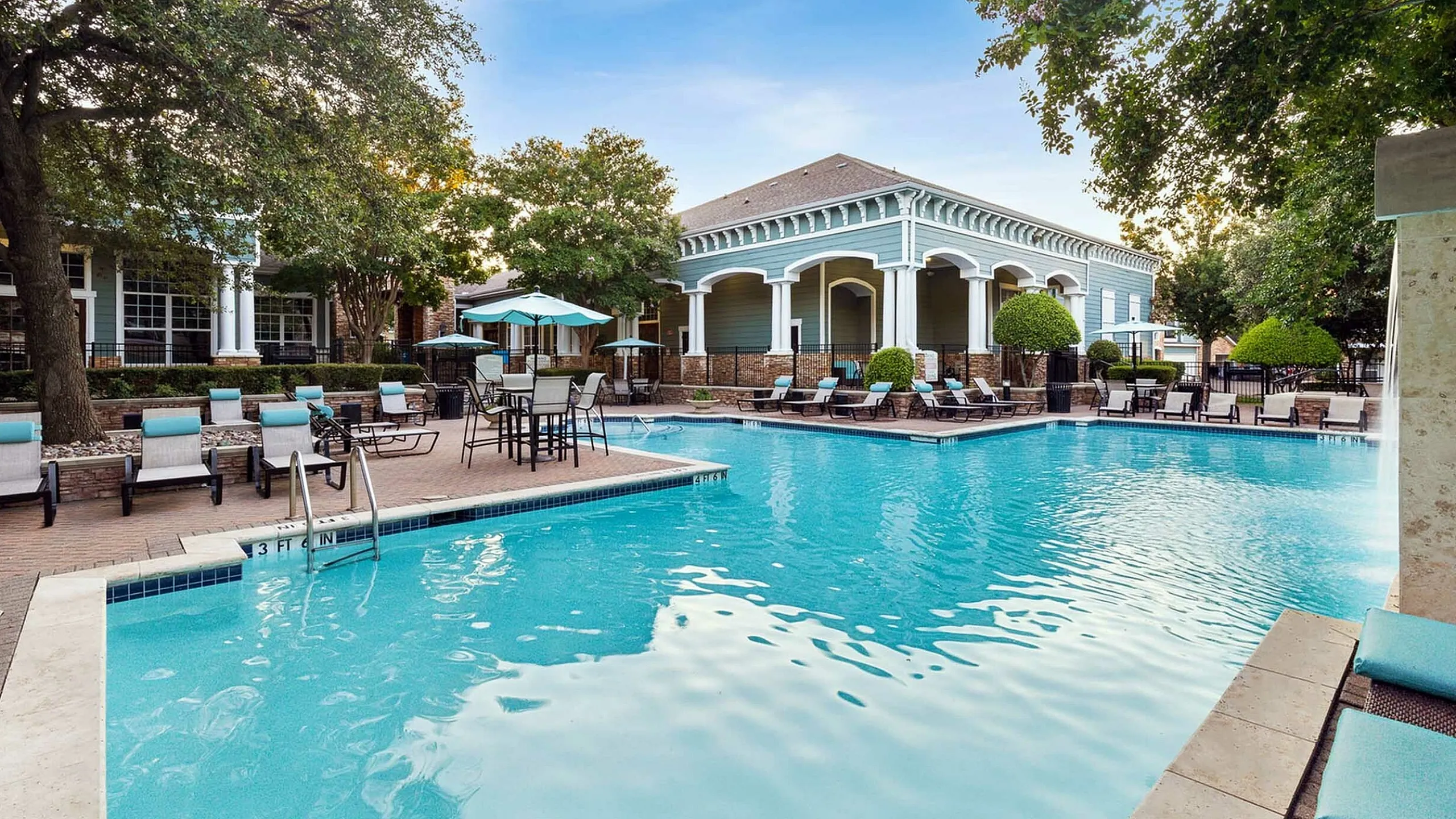 Luxurious apartment complex featuring a sparkling outdoor pool with sun loungers, shaded seating, and lush landscaping under a clear blue sky.