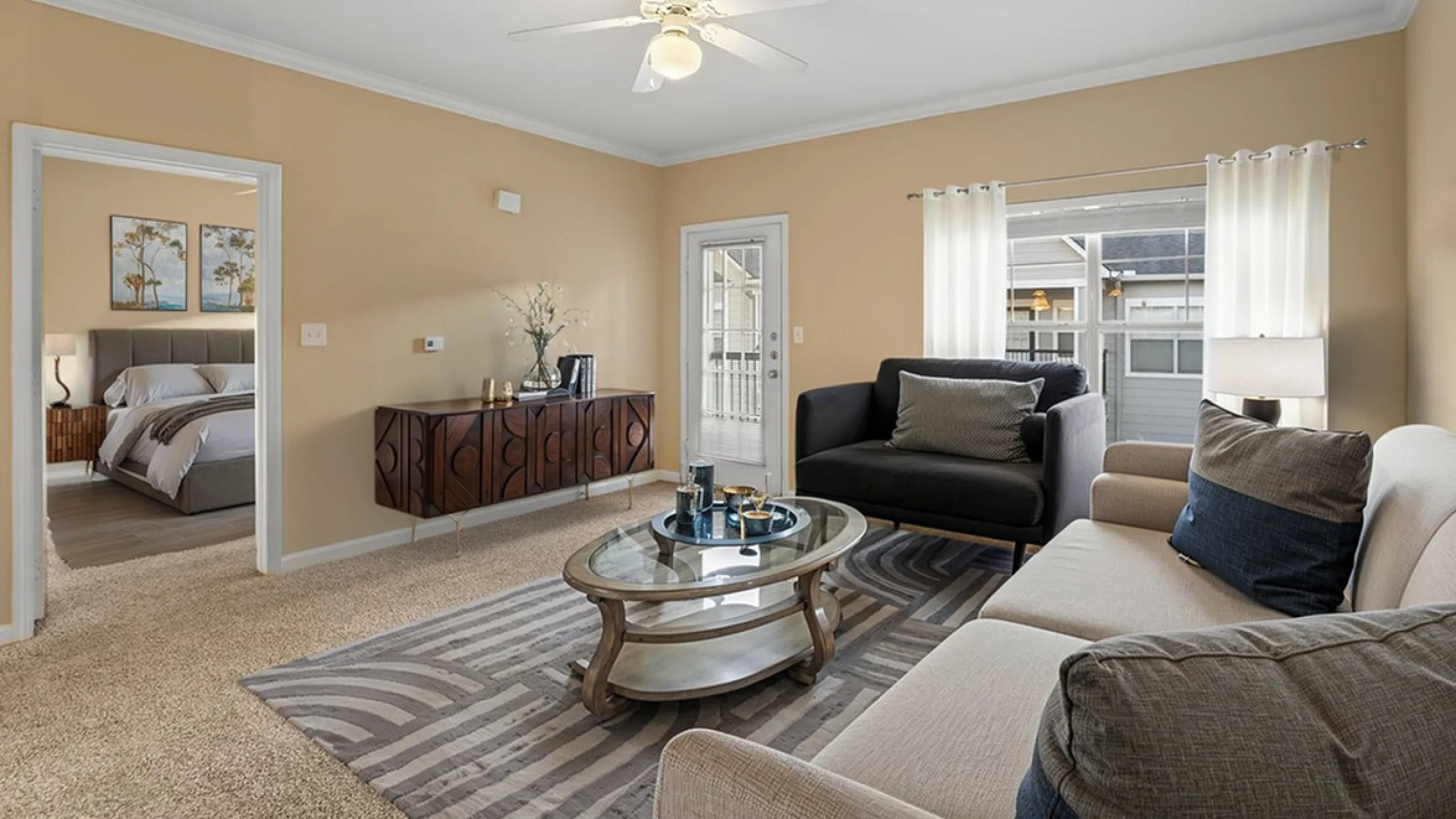 Elegant living room with plush sofas, a stylish glass coffee table, and contemporary decor, featuring a view into a cozy bedroom.