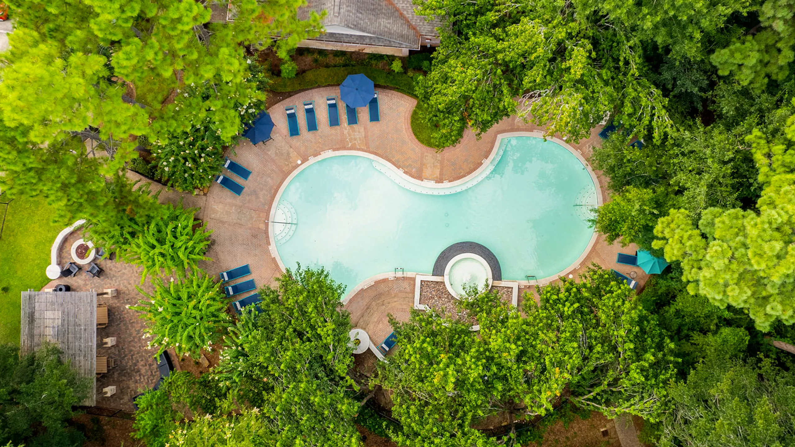 Aerial view of a luxurious, uniquely-shaped swimming pool surrounded by lush green trees and relaxing lounge chairs, offering a serene and private retreat at Raveneaux Apartments in Houston
