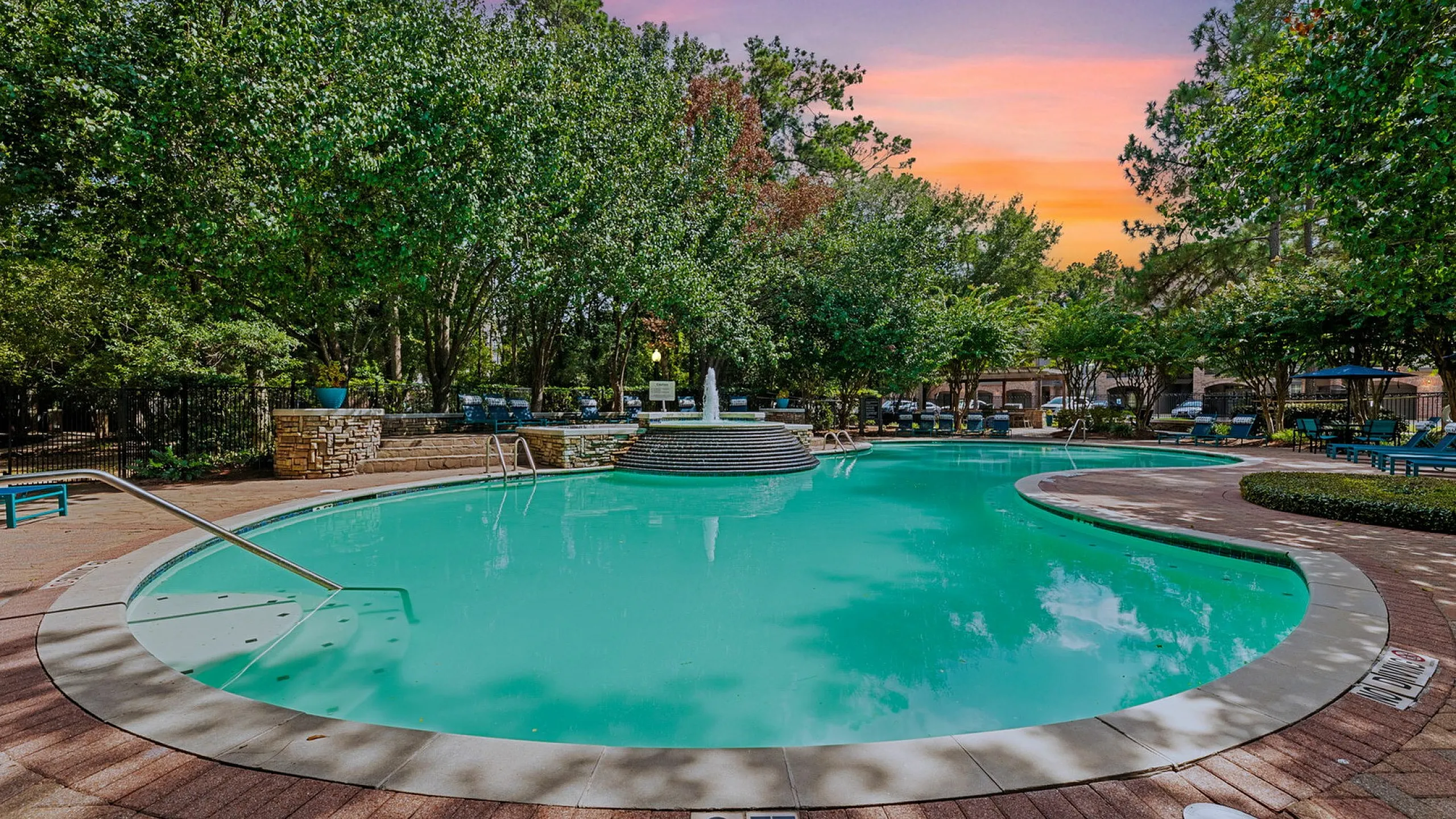 Inviting outdoor pool area surrounded by lush greenery, with a serene water fountain under a vibrant sunset sky.