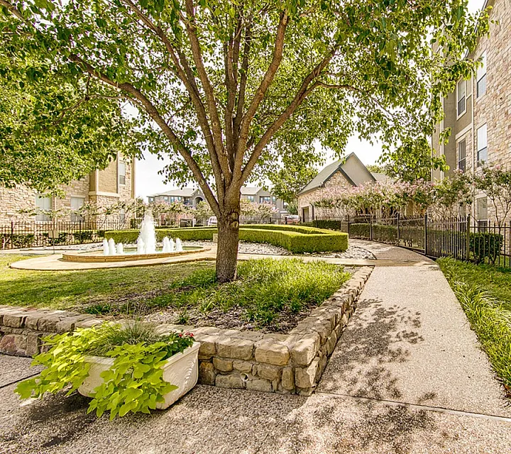 Charming apartment exterior with lush landscaping, vibrant greenery, and a serene fountain in a beautifully paved courtyard at Stonebriar at Frisco