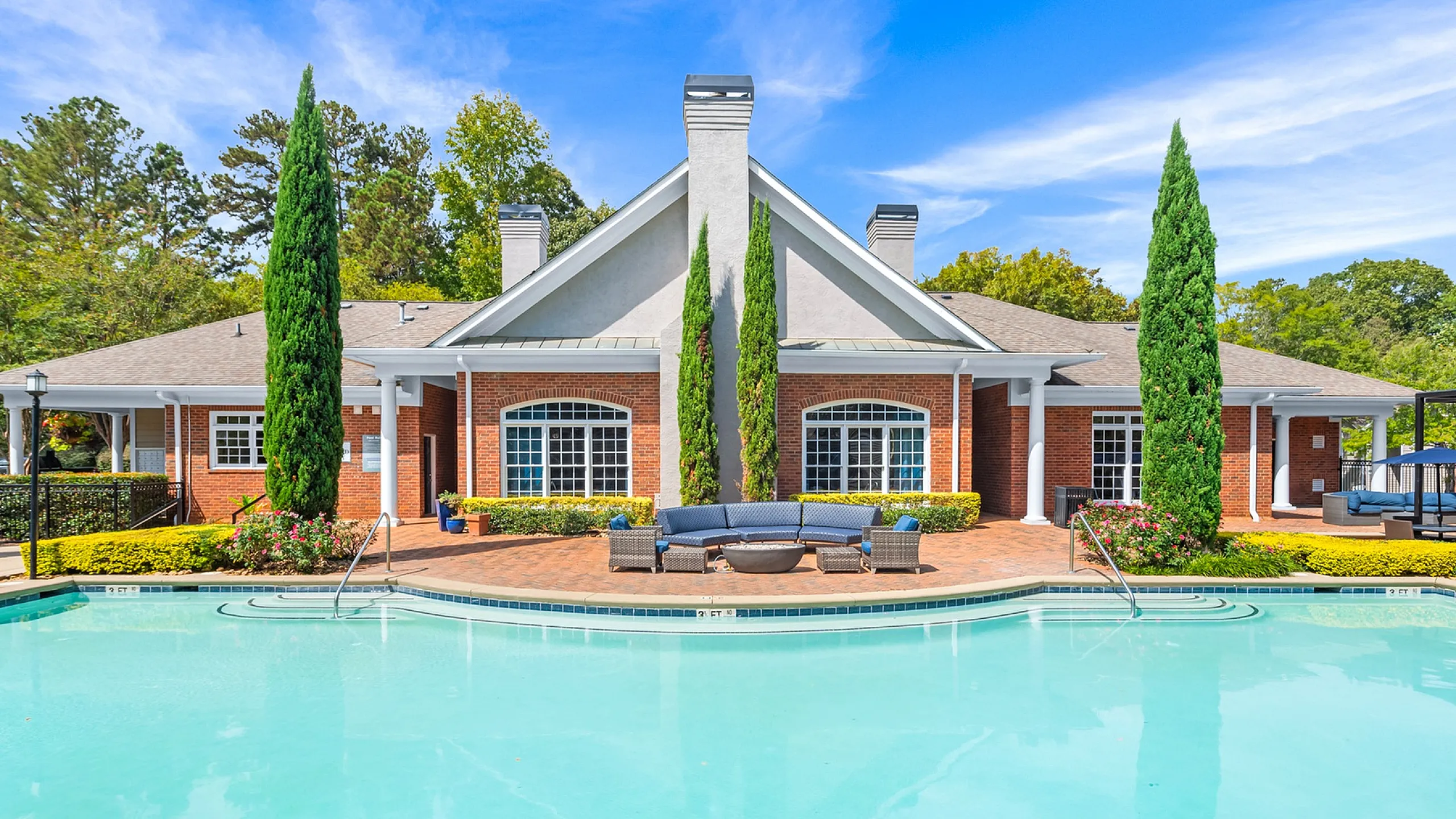 Luxurious community swimming pool with elegant brick clubhouse, stylish outdoor seating, and lush landscaping under a bright blue sky Whether you're enjoying a match on the tennis court or strolling on the on-site walking paths, outdoo