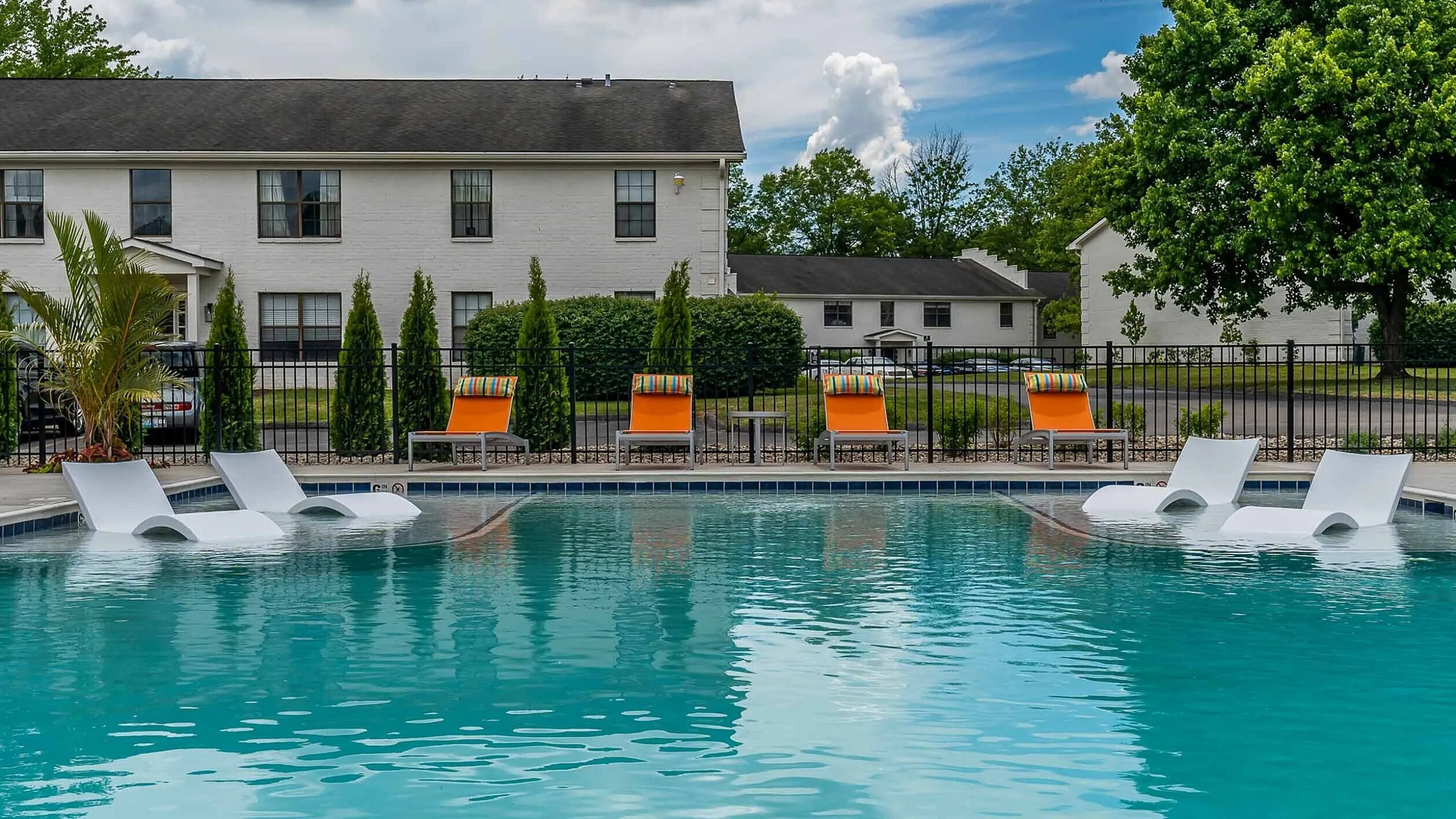 Inviting outdoor pool area with sleek white loungers and vibrant orange chairs, set against a backdrop of lush greenery and elegant apartments.