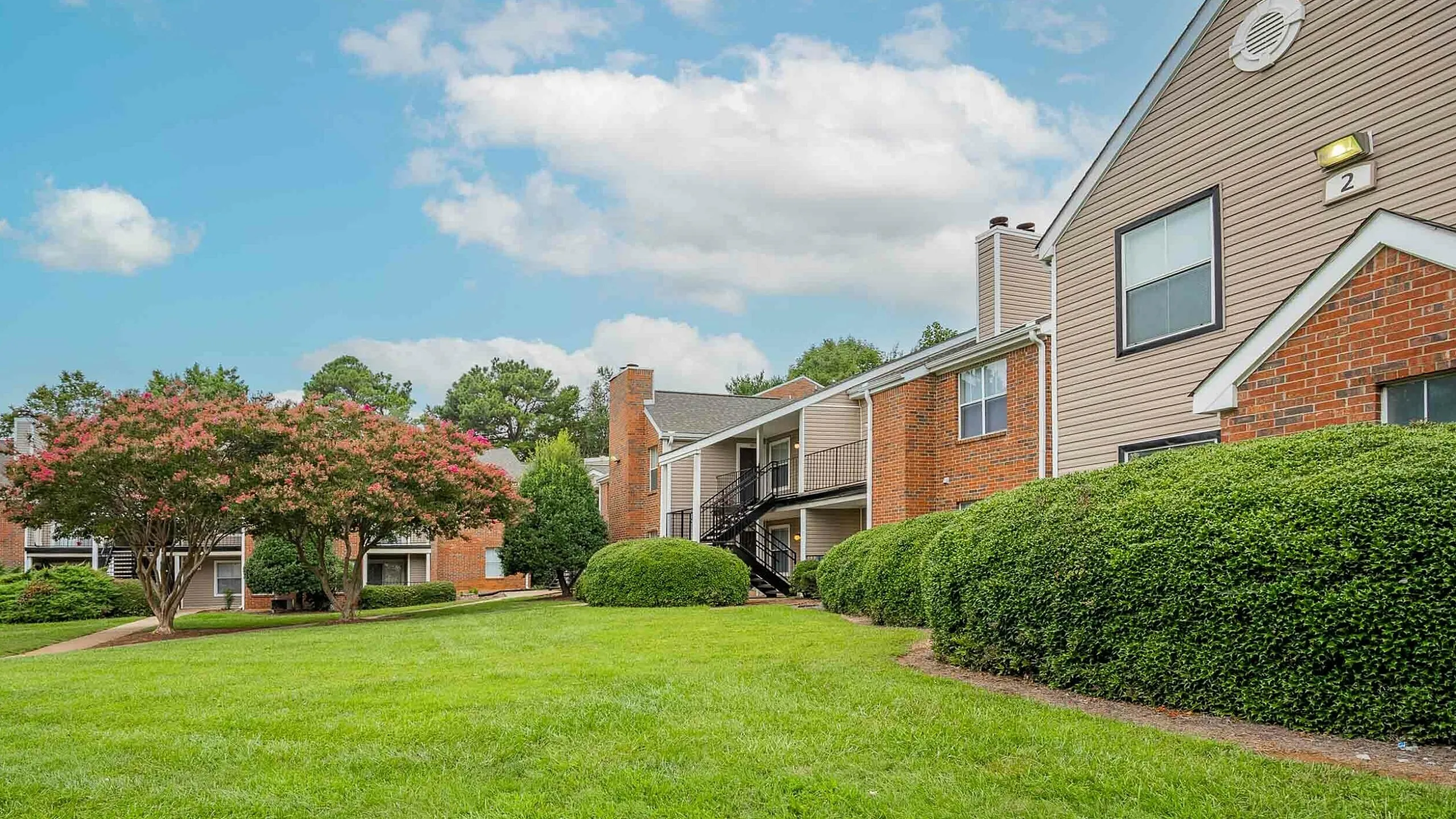 Charming apartment complex with manicured green lawns, beautiful flowering trees, and attractive brick exteriors under a clear blue sky.