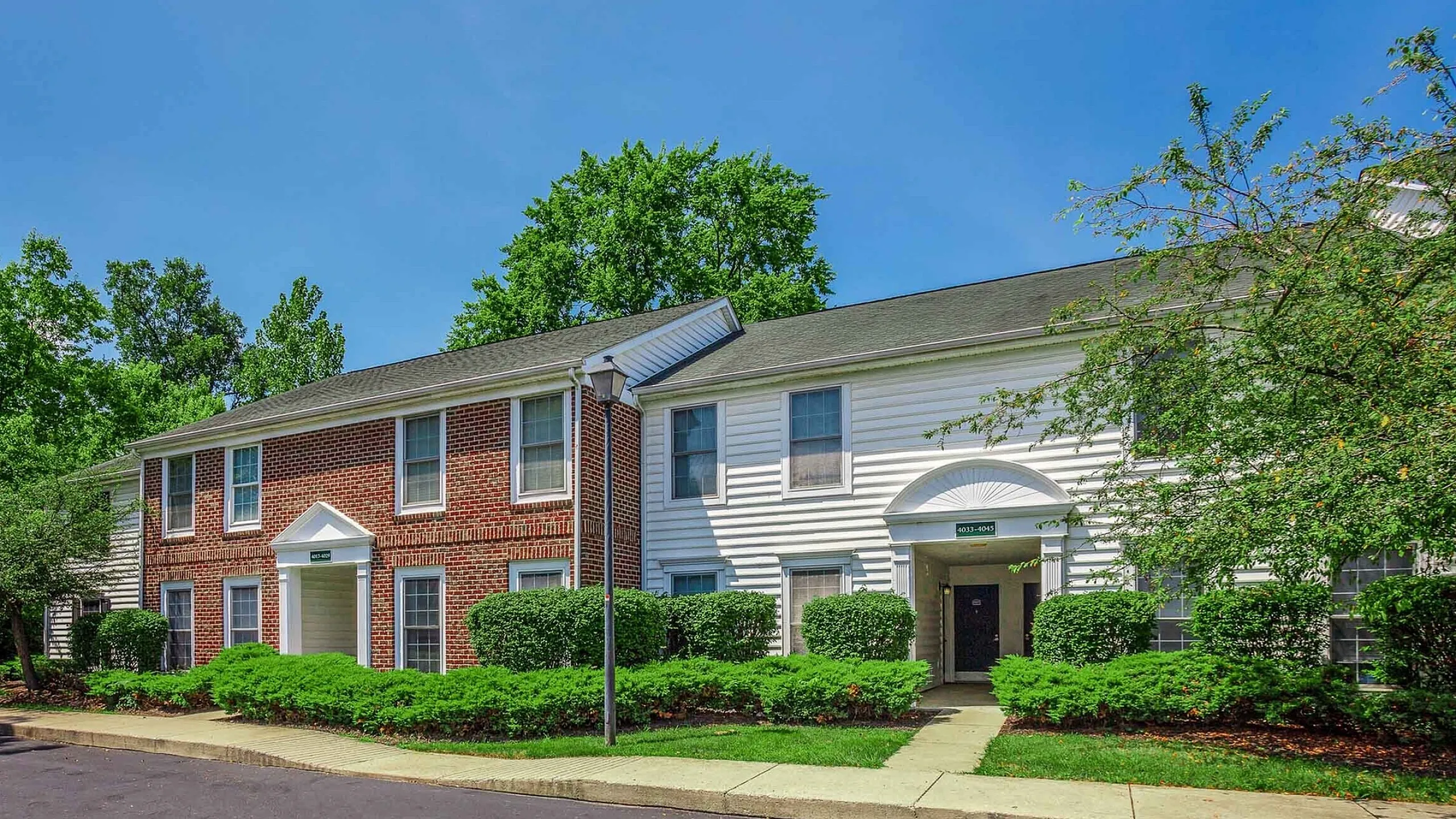 Charming two-story brick and siding apartment building surrounded by lush greenery and a clear blue sky.
