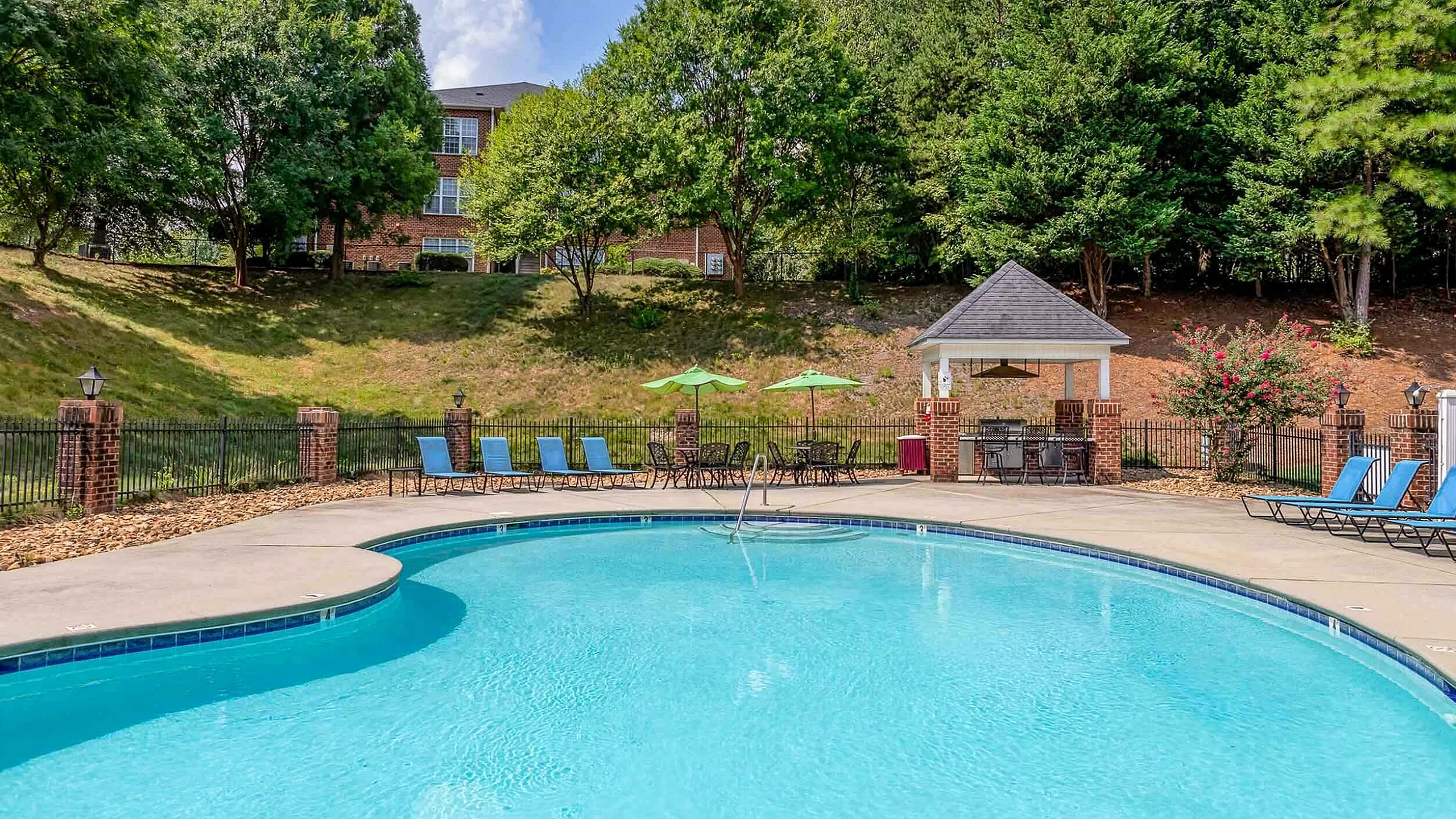 Inviting apartment complex pool area with vibrant blue water, surrounded by lush greenery, comfortable loungers, and charming gazebo for relaxation.