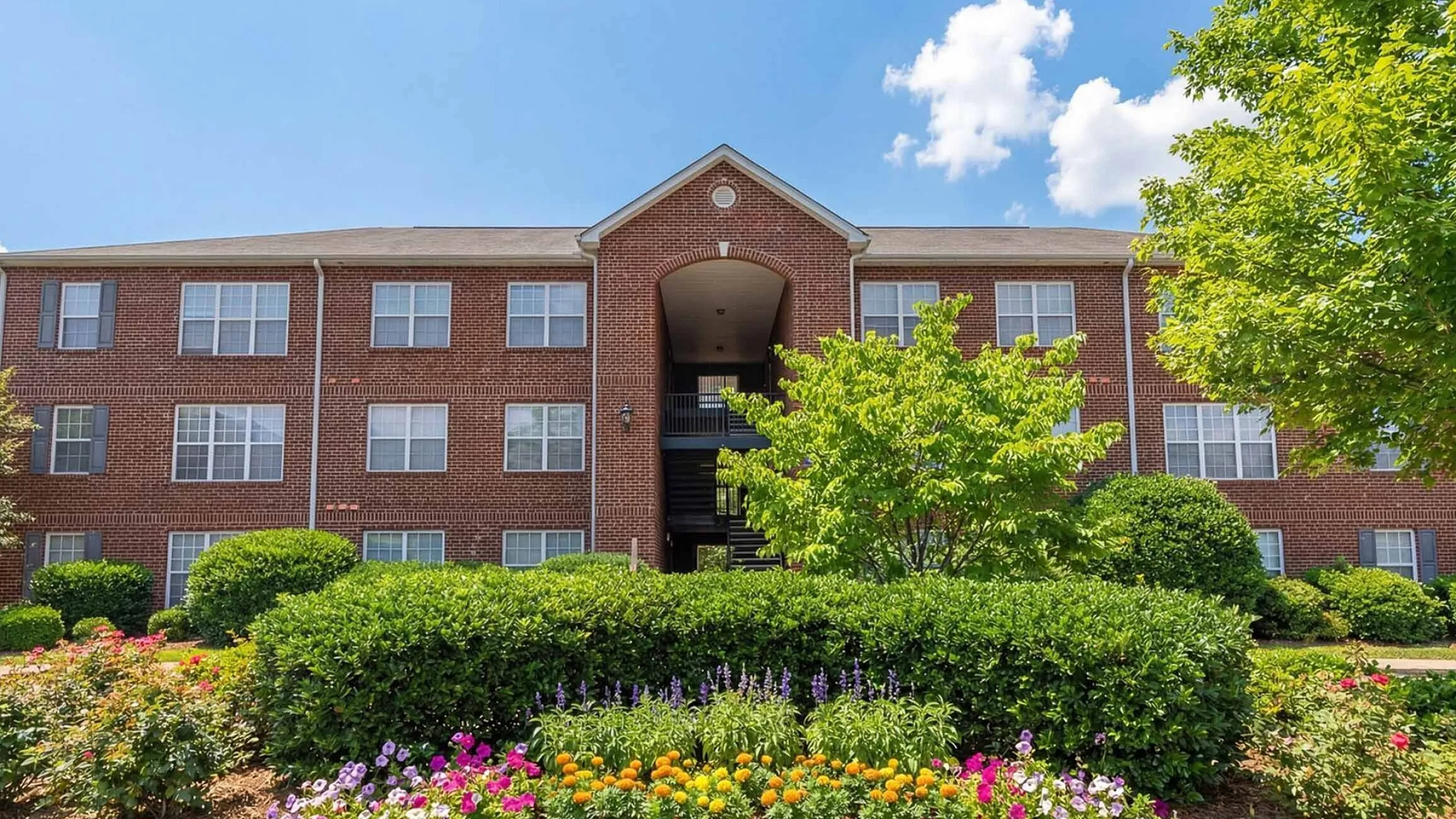 Charming red-brick apartment building with lush landscaping and vibrant flower beds under a bright blue sky.