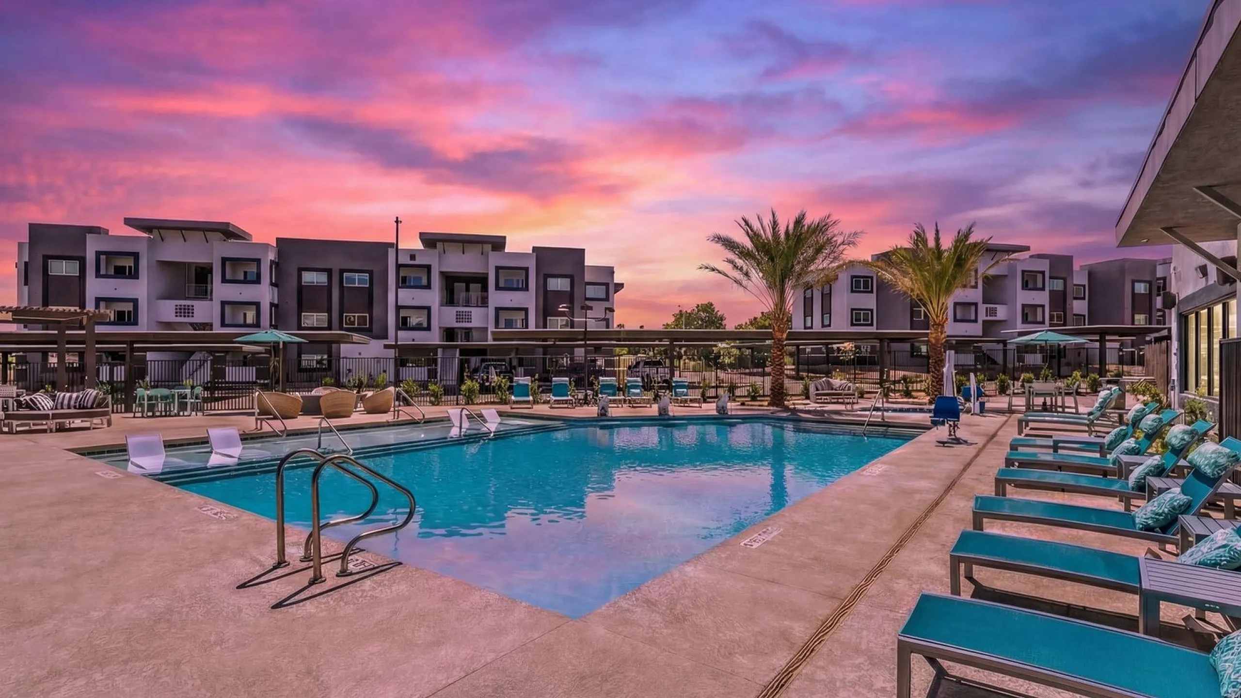 Luxurious apartment complex with a sparkling pool, surrounded by stylish lounge chairs and palm trees, set against a breathtaking sunset sky.