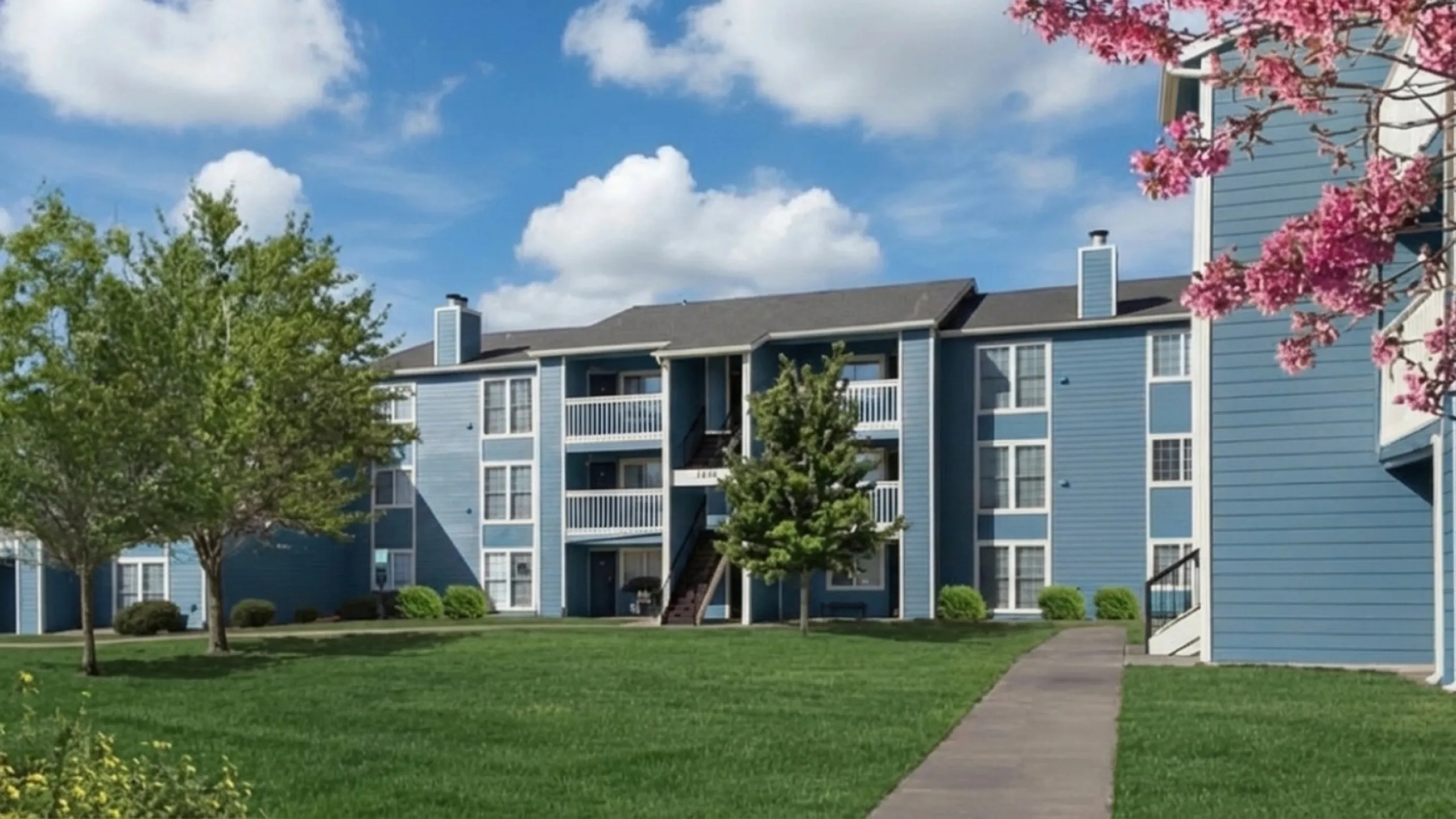 Beautiful apartment complex with a modern blue exterior set against a vibrant green lawn and blooming pink flowers under a blue sky.