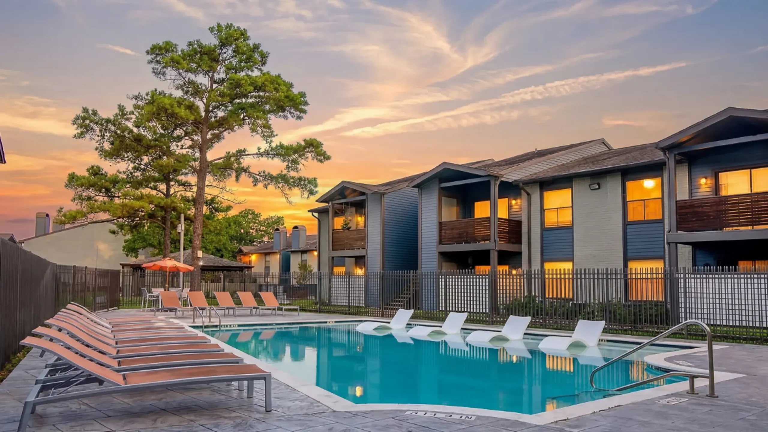 Inviting outdoor pool area surrounded by stylish apartment buildings, featuring ample lounge seating and scenic tree views at dusk.