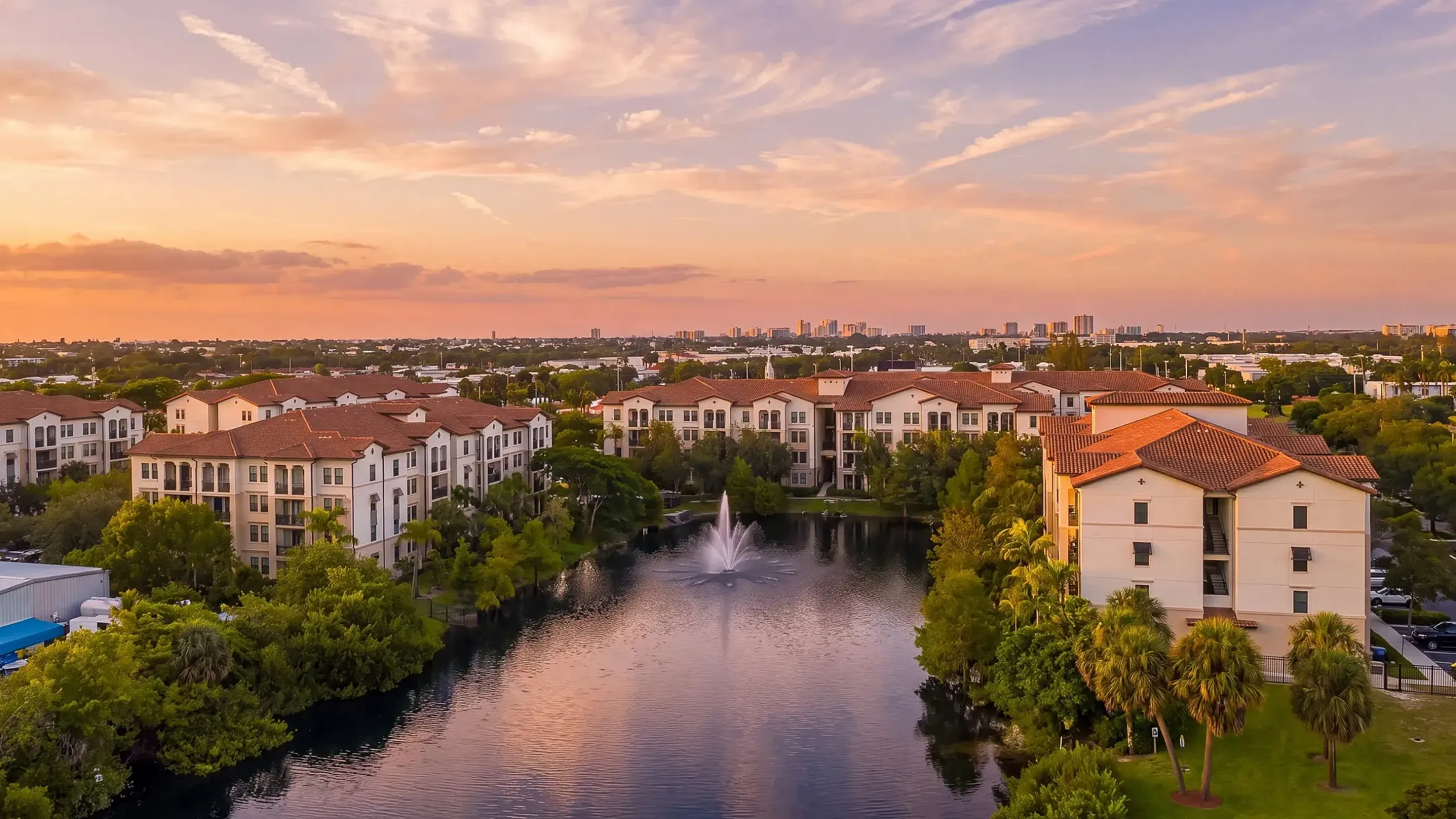 Stunning aerial view of a luxury apartment community with elegant architecture, a serene lake with a fountain, and a vibrant sunset sky.