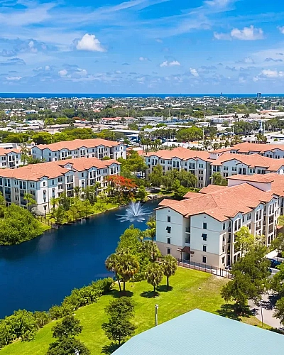 Aerial view of a vibrant apartment complex with terracotta roofs, surrounded by lush greenery and a tranquil water feature, set against a clear blue sky backdrop.