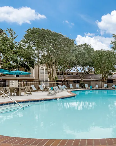 Gorgeous apartment complex pool with a clear blue water, surrounded by comfortable lounge chairs and lush green trees under a bright sunny sky.