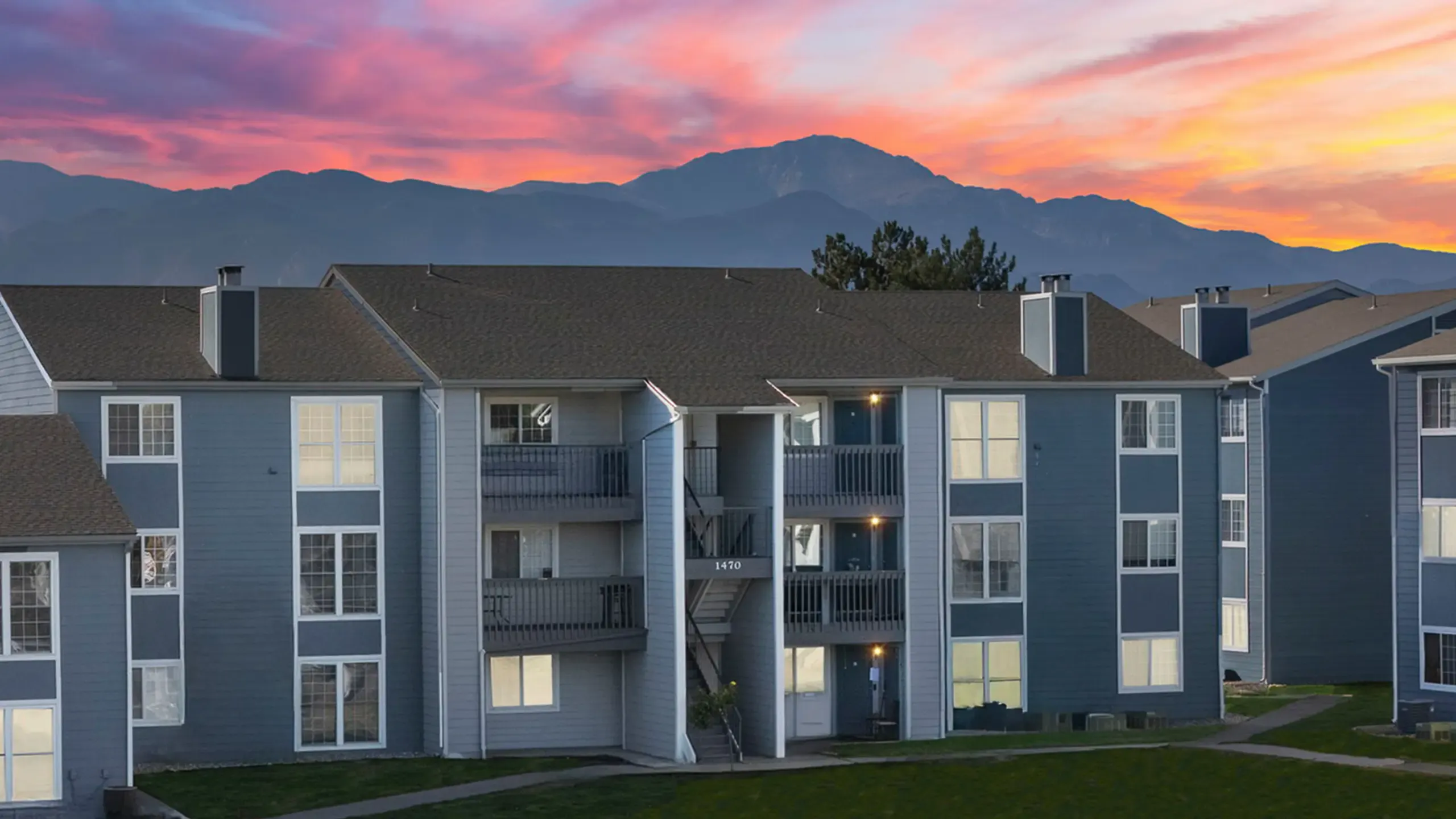 Beautiful apartment complex with a modern blue exterior set against a vibrant green lawn and blooming pink flowers under a blue sky.