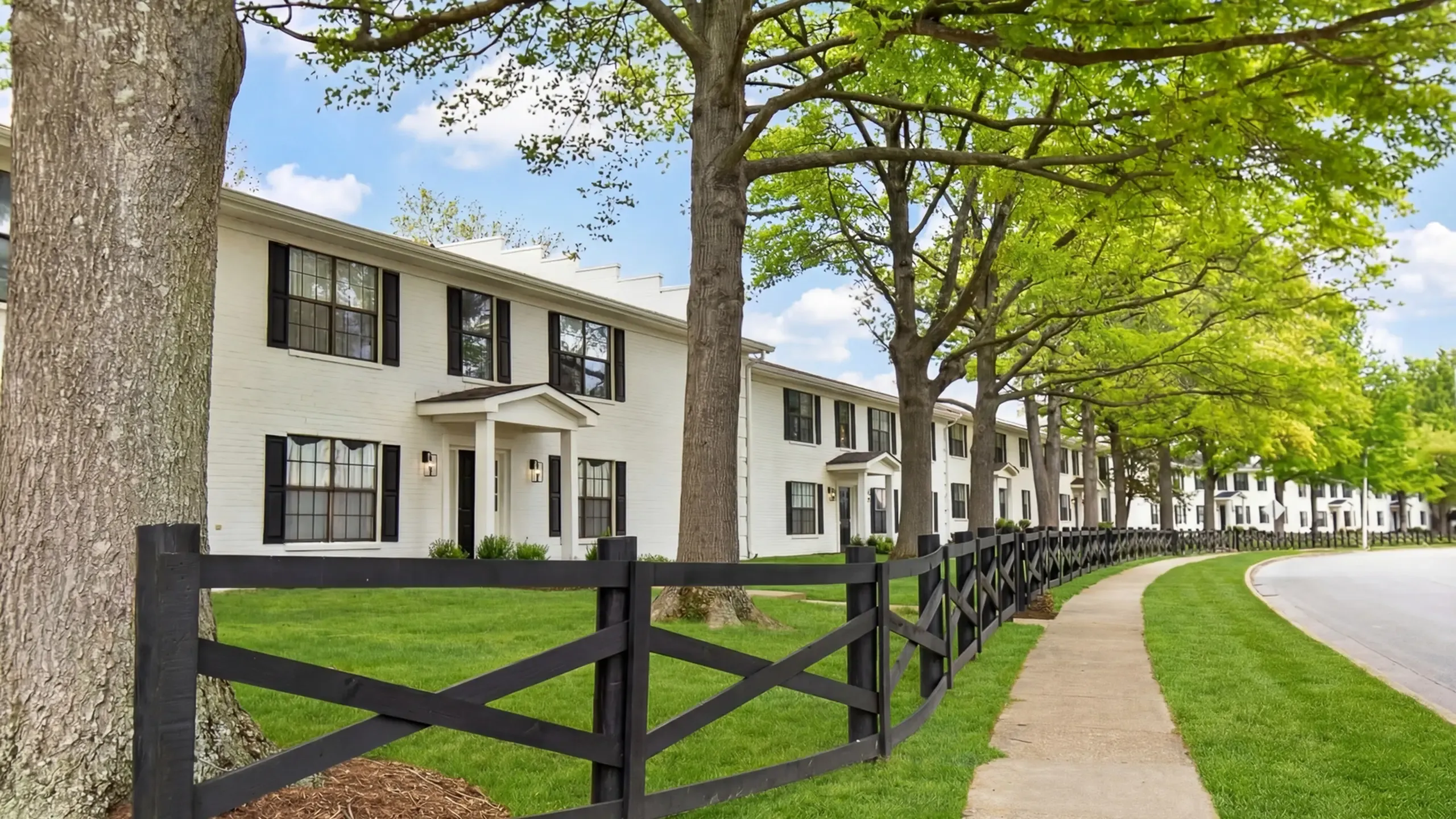 Charming residential area featuring classic white townhouses with black trim, set along a tree-lined street with manicured lawns and a scenic walkway.