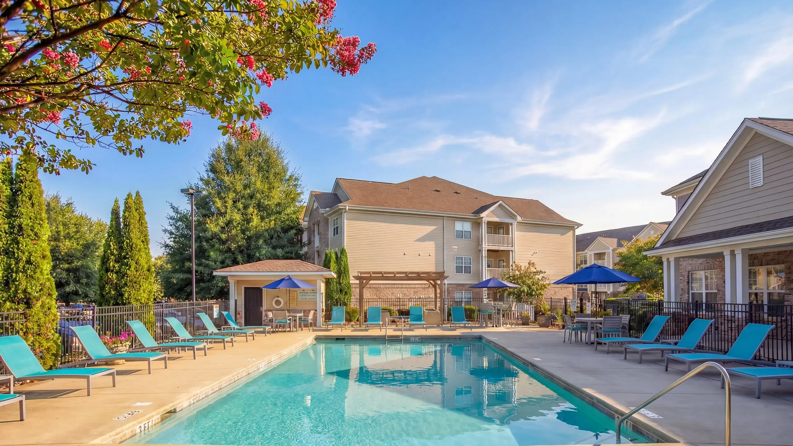 Inviting outdoor pool area with vibrant blue lounge chairs, surrounded by lush greenery and charming apartment buildings at Stratford at Hillcrest Apartments in Winston-Salem.