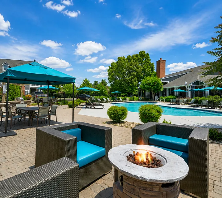 Luxurious outdoor patio with cozy seating, a firepit, and a sparkling pool surrounded by lush greenery and shaded umbrellas.