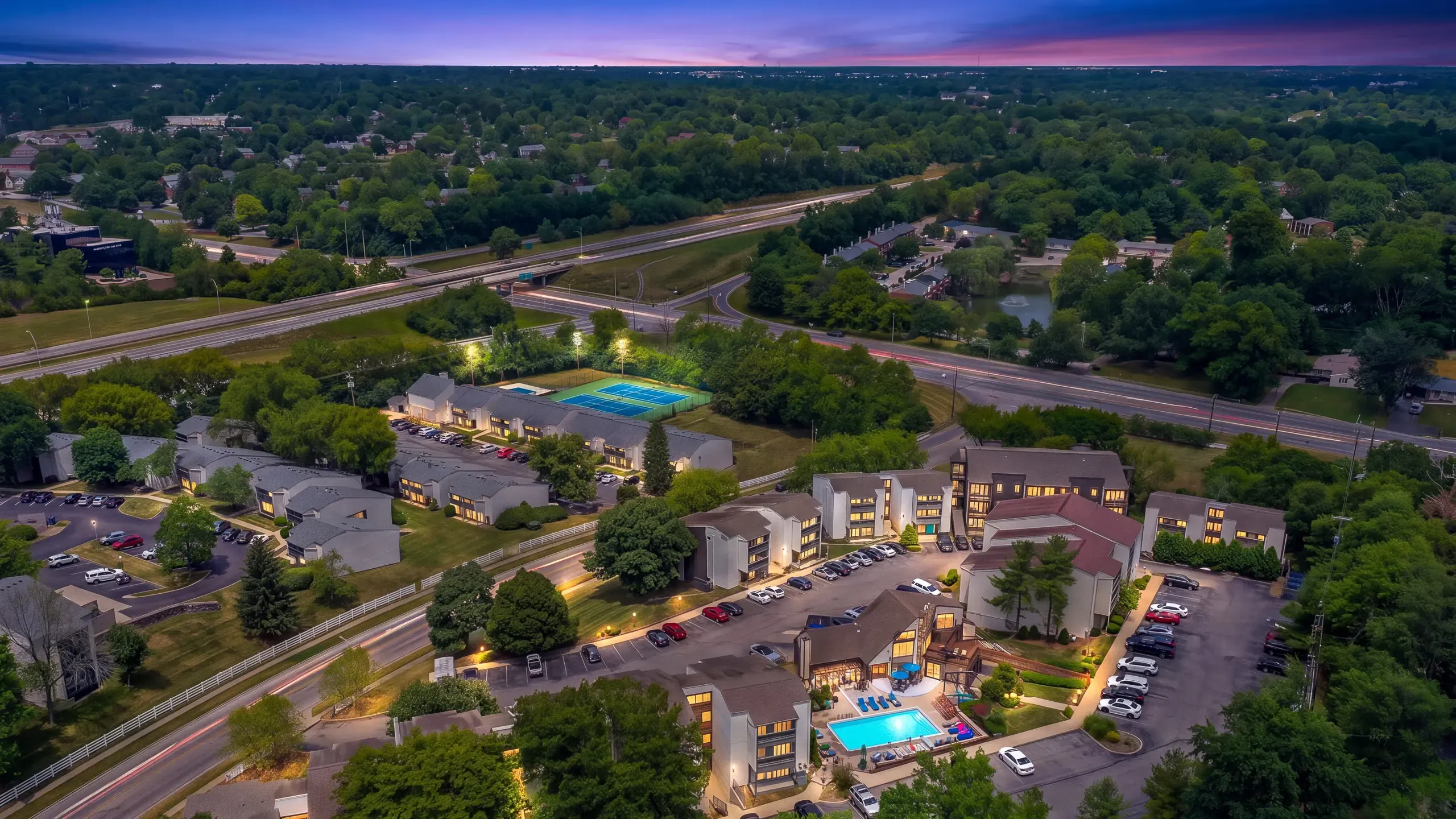 Aerial view of a luxury apartment complex featuring illuminated swimming pool, vibrant green landscapes, and modern architecture at dusk.