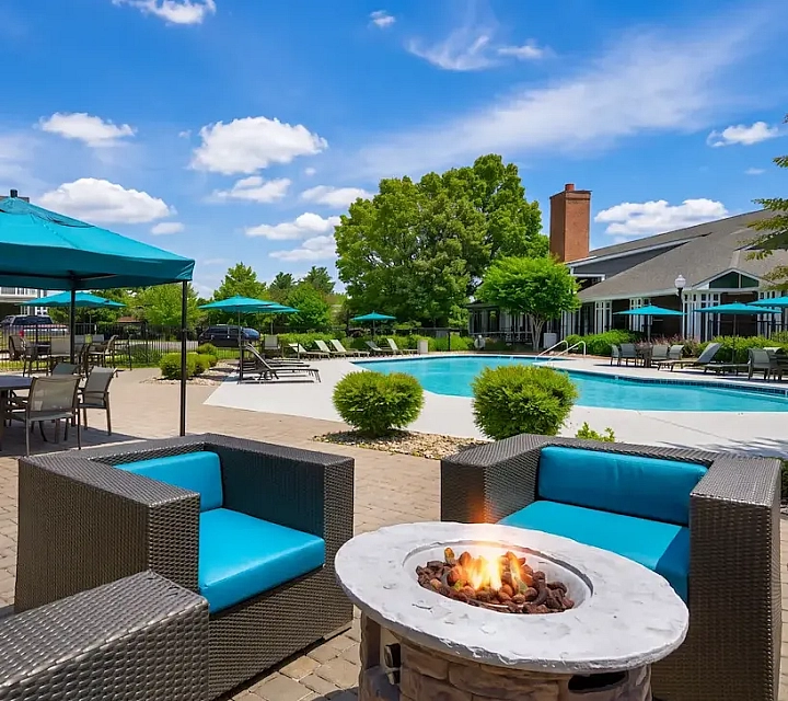 Luxurious outdoor patio with cozy seating, a firepit, and a sparkling pool surrounded by lush greenery and shaded umbrellas.