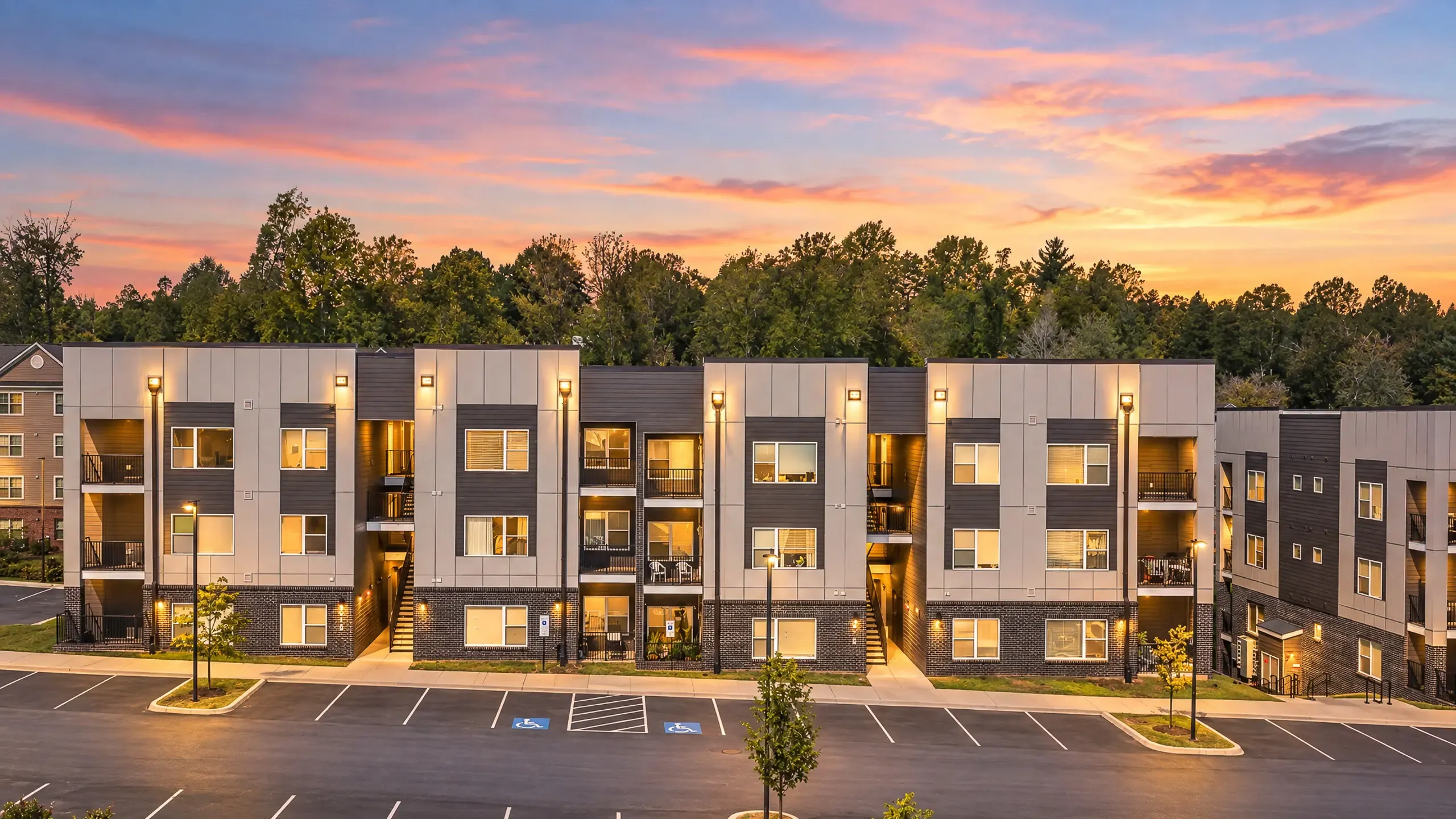 Modern apartment complex at sunset, showcasing sleek architecture with balconies and ample parking, surrounded by lush greenery.