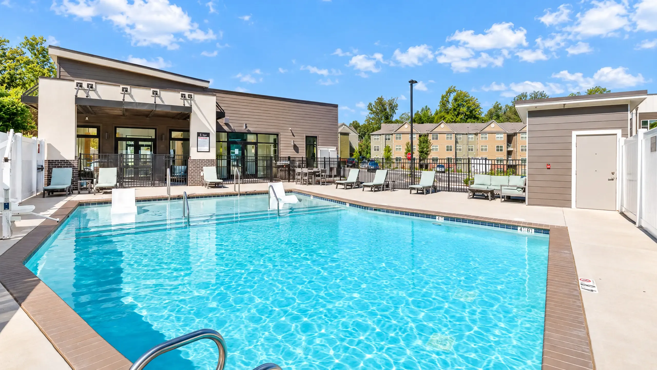 Inviting outdoor pool area with lounge chairs and modern clubhouse under a bright blue sky, perfect for relaxation and leisure.