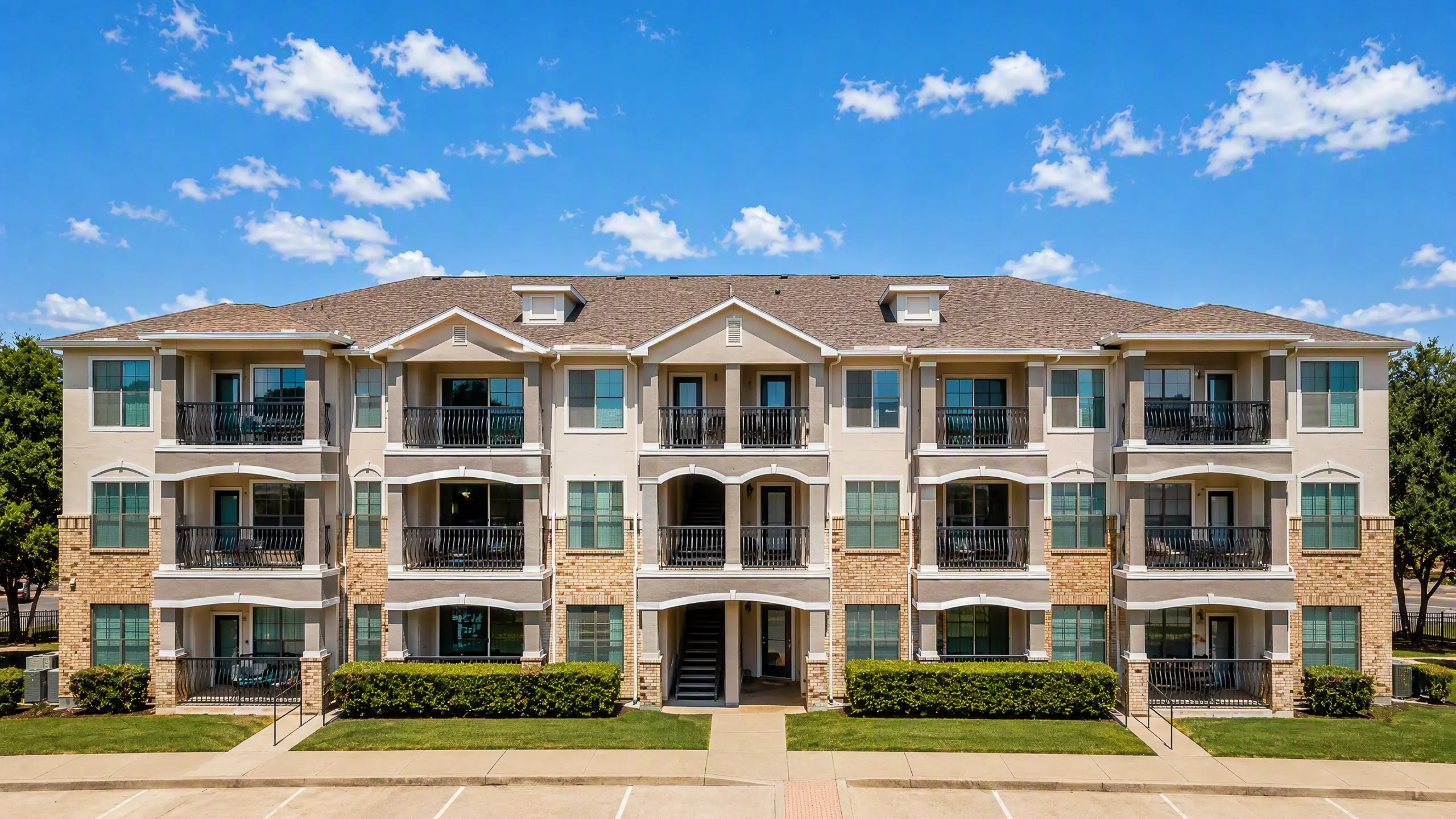 Inviting three-story apartment building with spacious balconies, surrounded by lush greenery and under a clear blue sky.