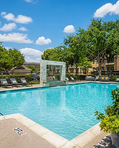Luxurious apartment community pool with comfortable lounge chairs, surrounded by lush greenery and bright blue skies.
