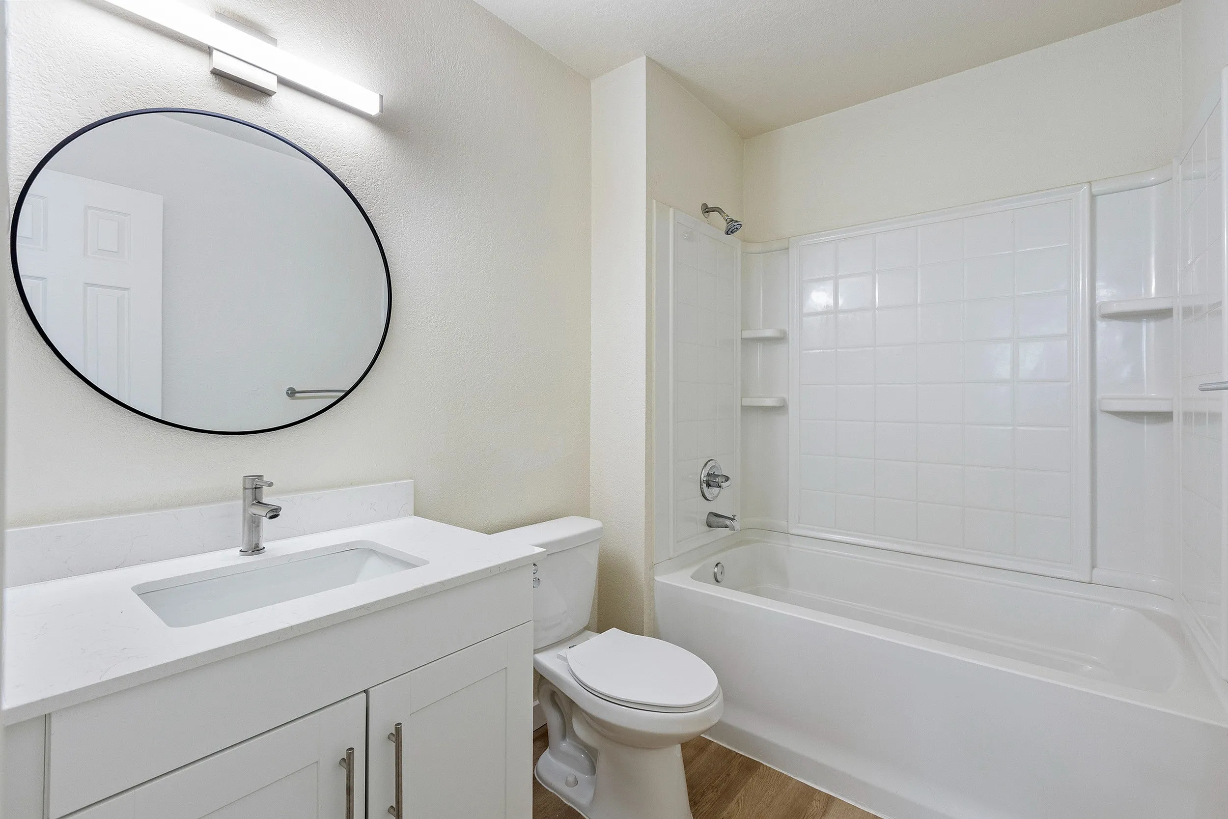 Modern bathroom featuring a white vanity, round mirror, chic hardwood flooring, and bathtub with elegant fixtures.