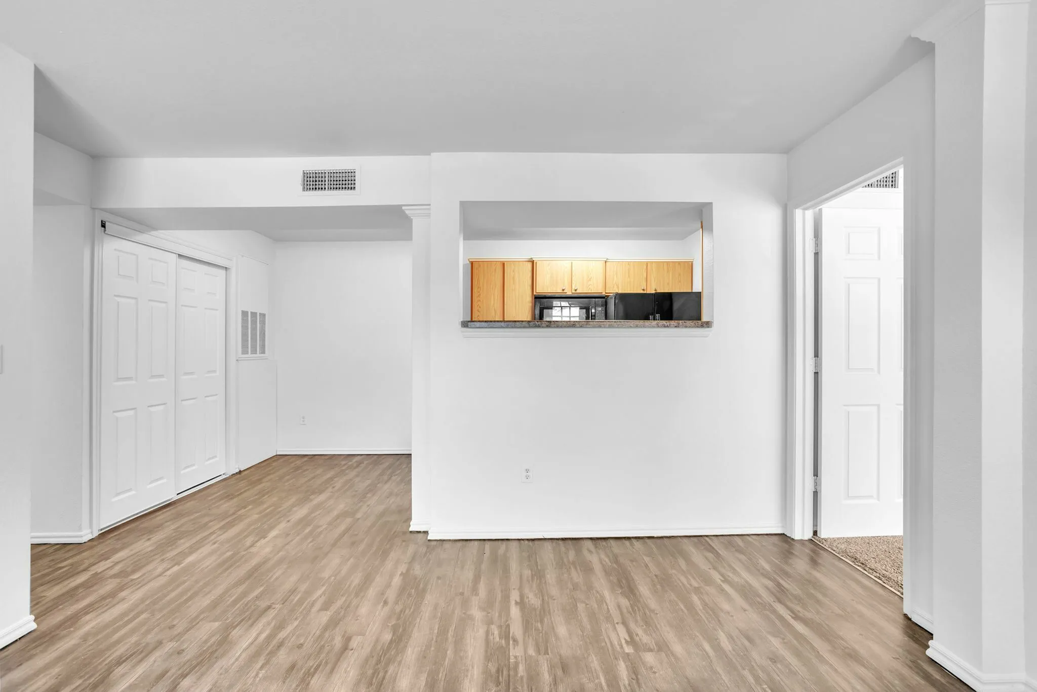 Bright apartment interior with open-concept layout, featuring a modern kitchen with wooden cabinets and sleek appliances against light-colored walls and flooring.
