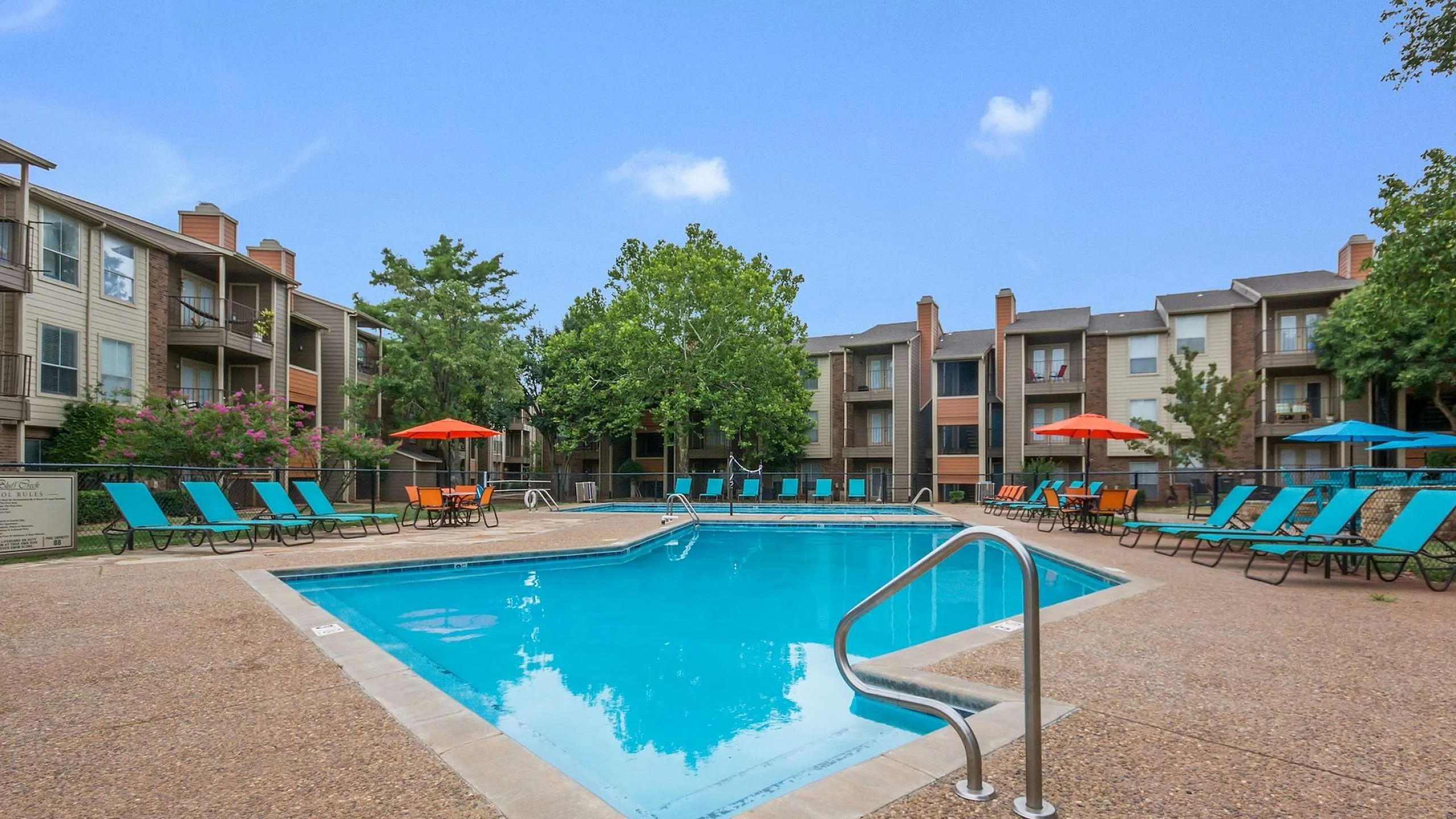 Bluff Creek Apartment complex swimming pool with clear blue water, surrounded by teal-cushioned lounge chairs, orange umbrellas, and a volleyball net, with brick buildings and trees in the background in Northwest OKC.
