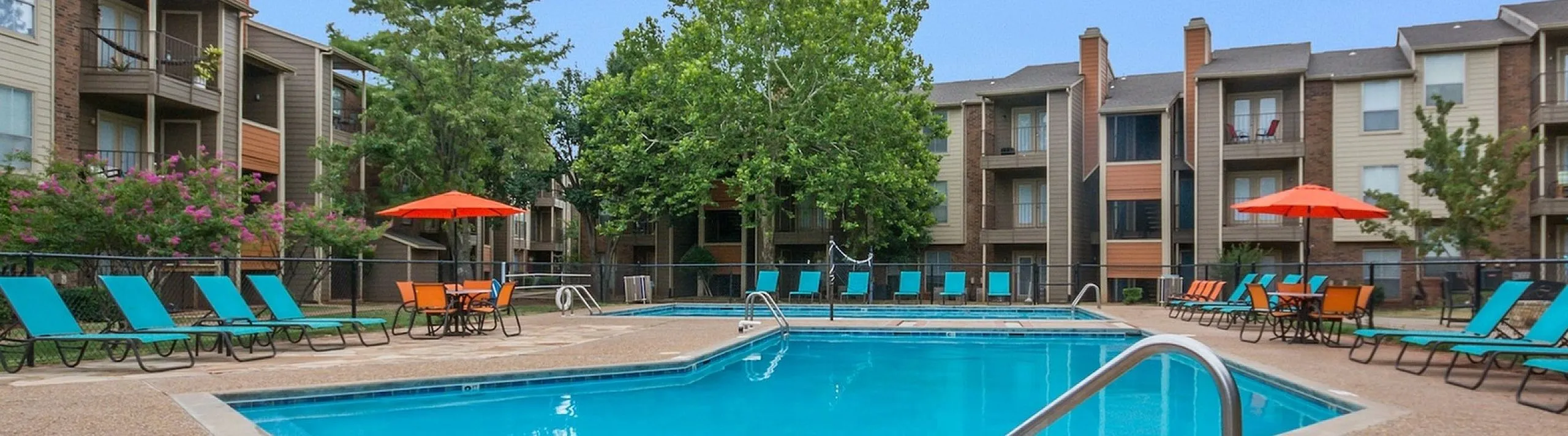 Apartment complex swimming pool with clear blue water, surrounded by teal-cushioned lounge chairs, orange umbrellas, and a volleyball net, with brick buildings and trees in the background in NW OKC