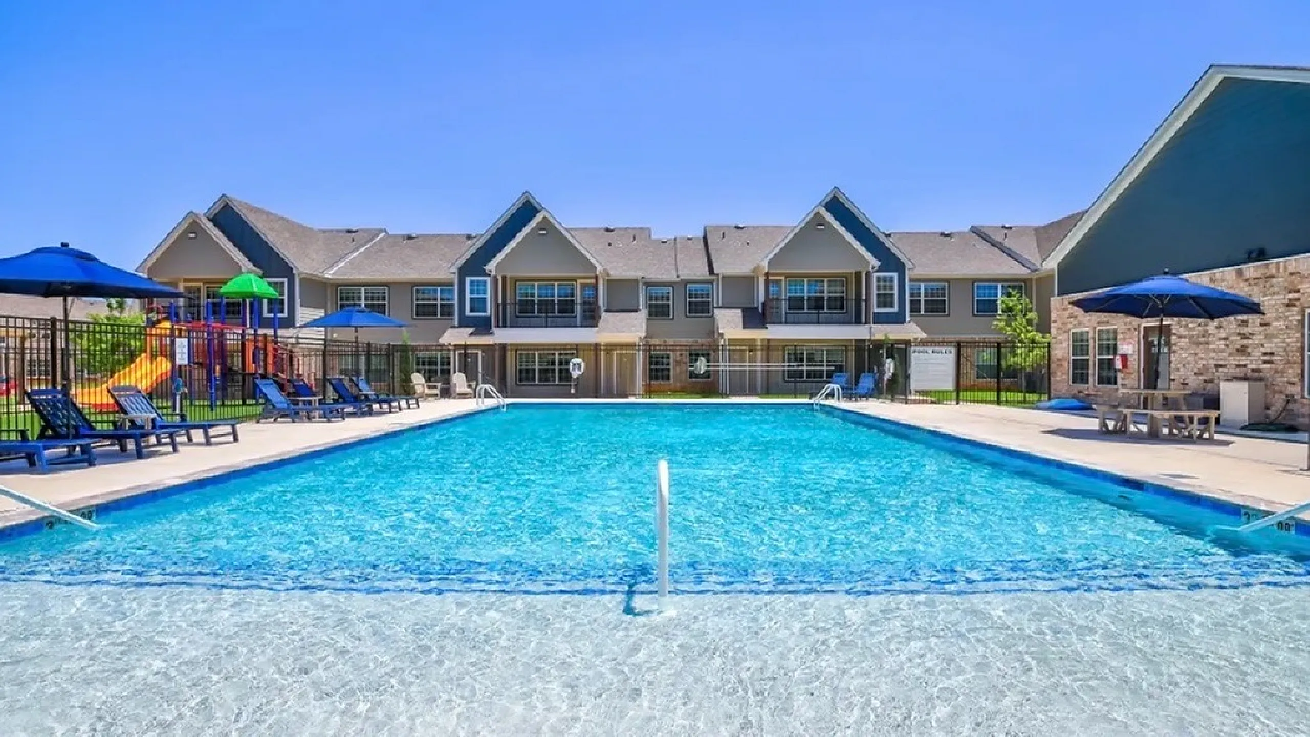 Vibrant apartment complex featuring a sparkling swimming pool, surrounded by cozy lounge chairs and colorful umbrellas under a clear blue sky.