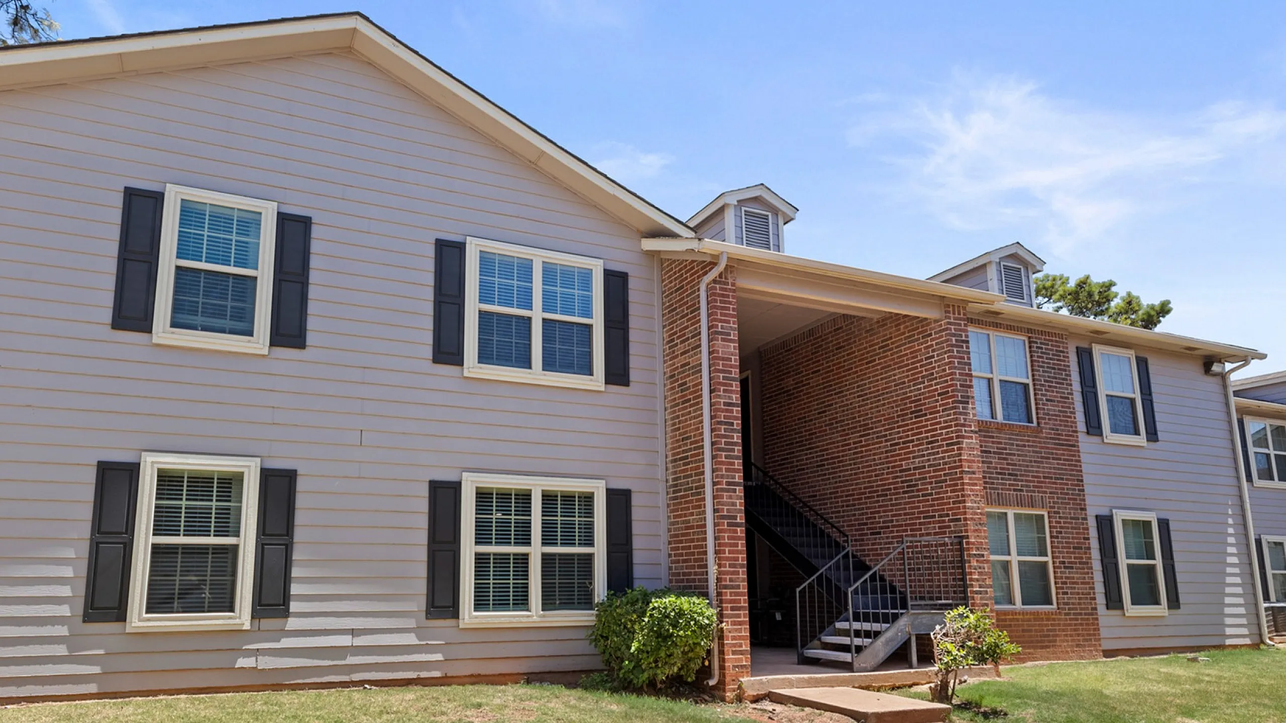 Charming two-story apartment building with brick and siding exterior, nestled in a landscaped setting under a clear blue sky.