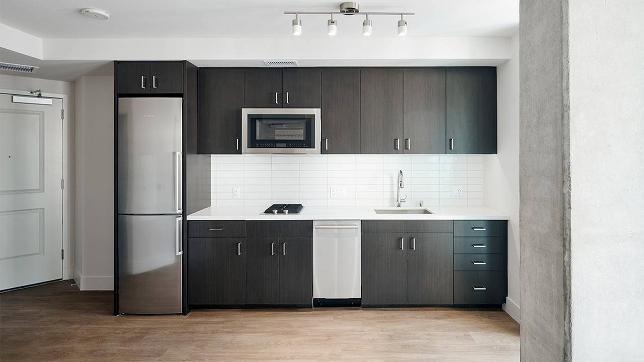 Dark cabinetry is accented by white quartz countertops and tile backsplash with silver stainless steel appliances, single door compact fridge and concrete accent wall