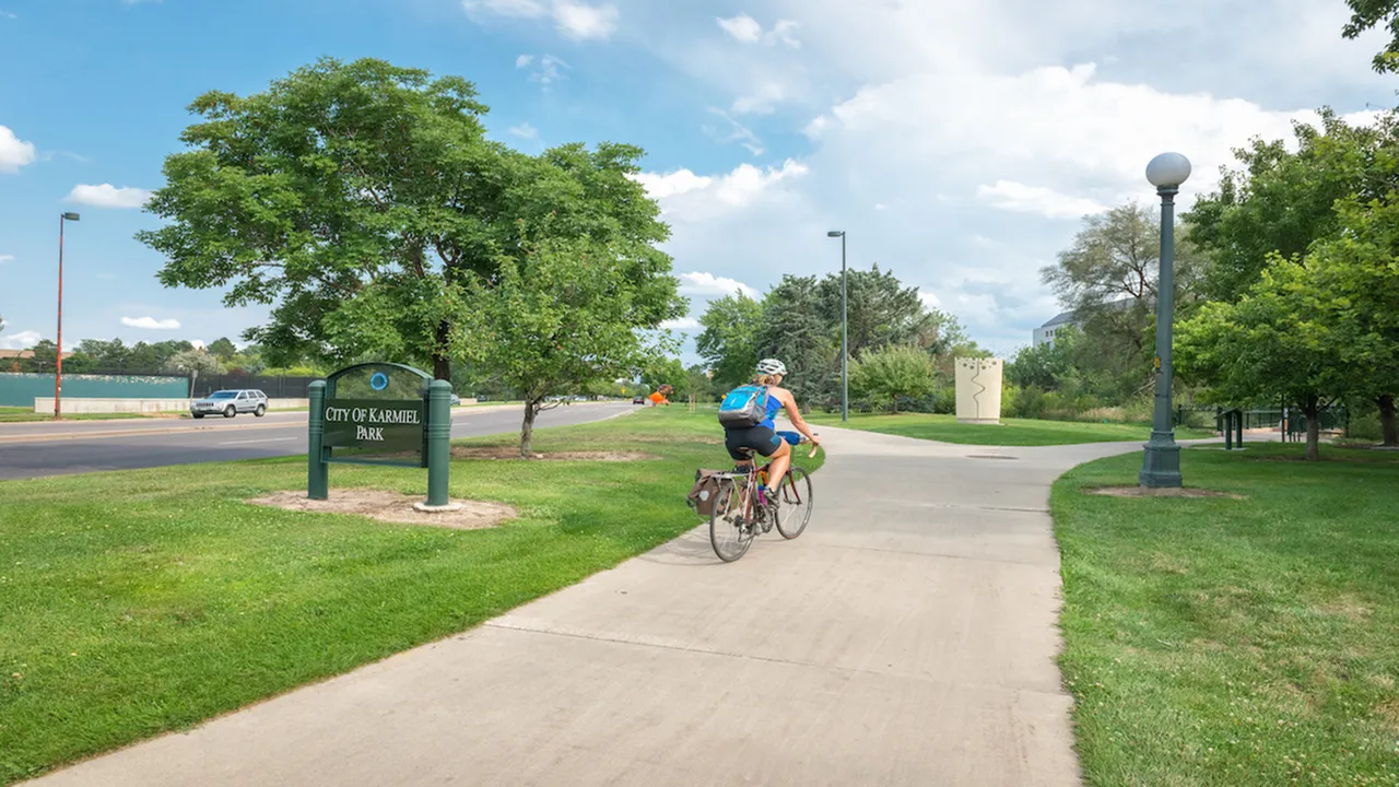 Scenic park pathway with a cyclist enjoying a sunny day at City of Karmiel Park, surrounded by lush greenery and clear blue skies.
