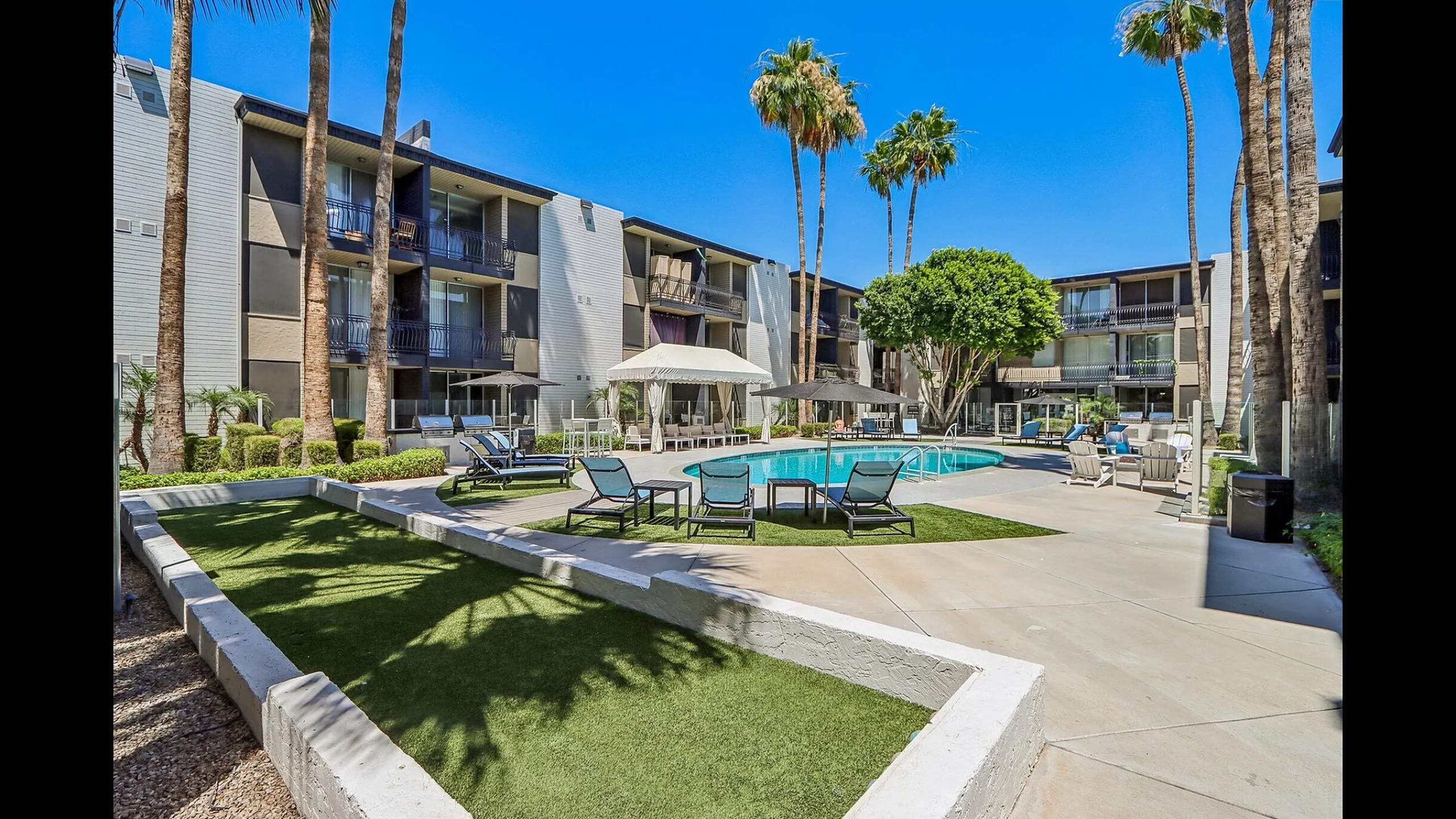 Chic apartment courtyard with a sparkling pool, lounge chairs, and lush palm trees under a bright blue sky.