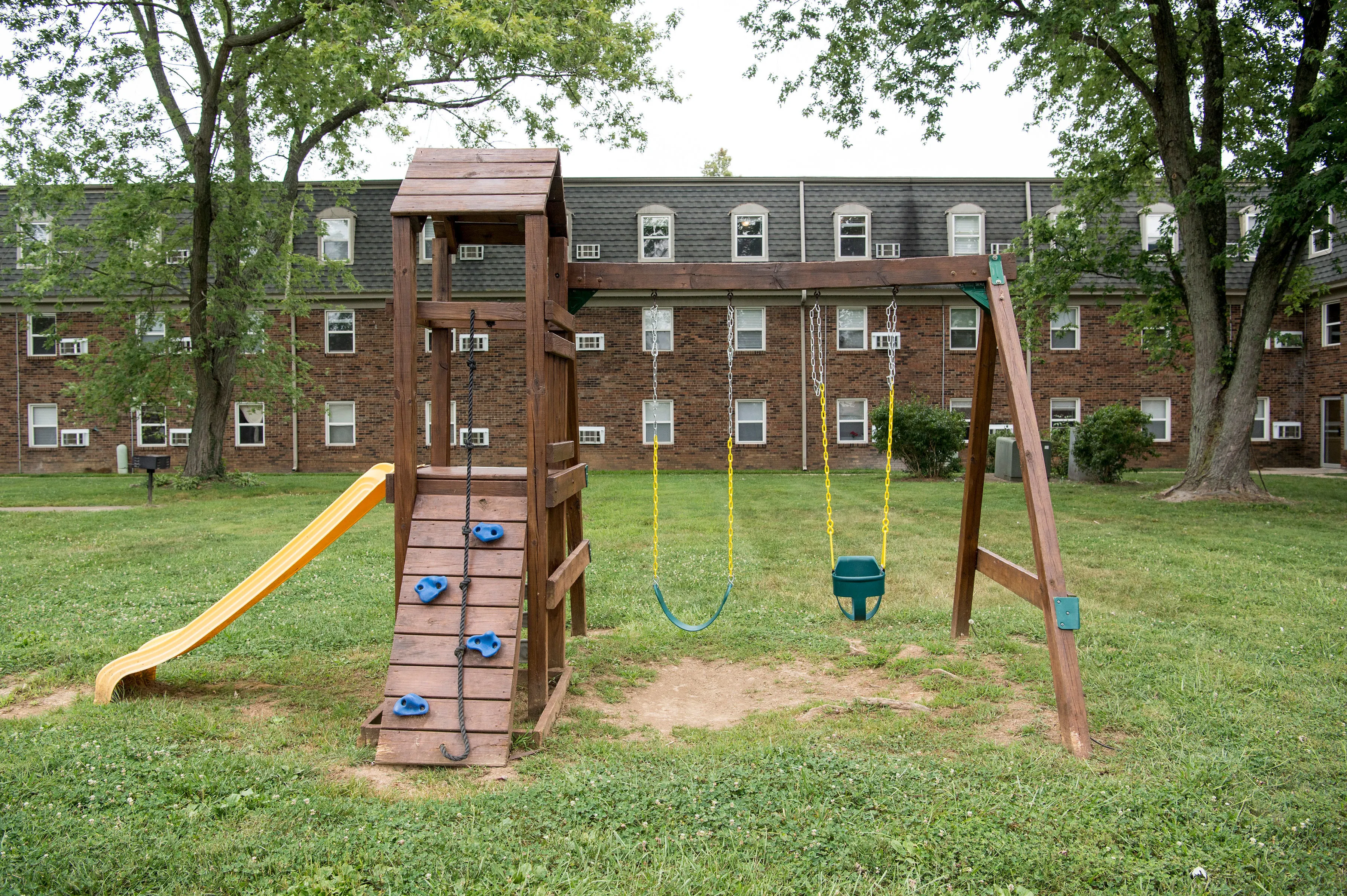 Charming residential playground with a wooden slide, swings, and climbing wall, situated in a lush green courtyard of a brick apartment building.