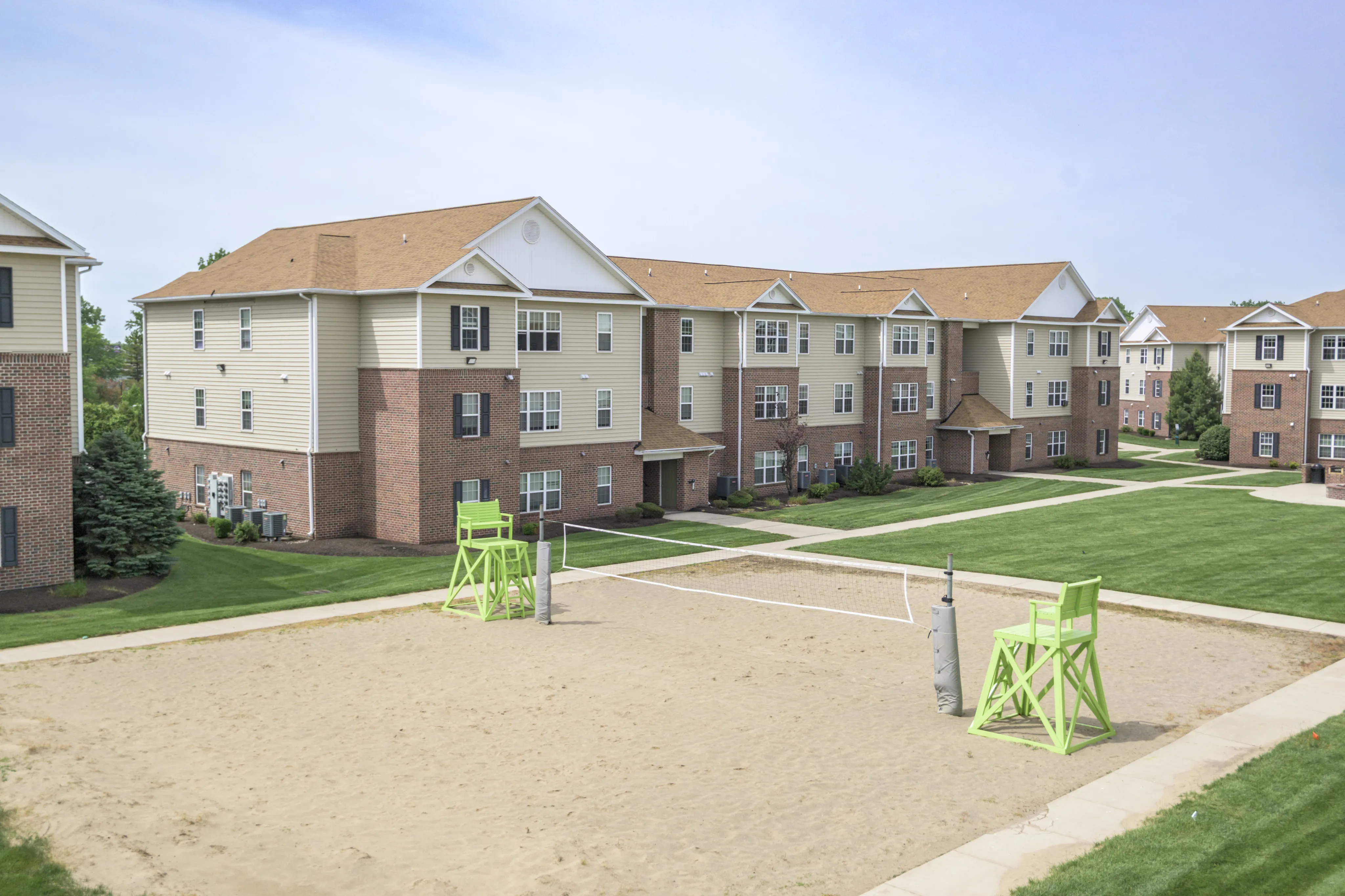 Inviting apartment complex with charming brick exterior, lush green lawns, and a recreational sand volleyball court, perfect for vibrant community living.