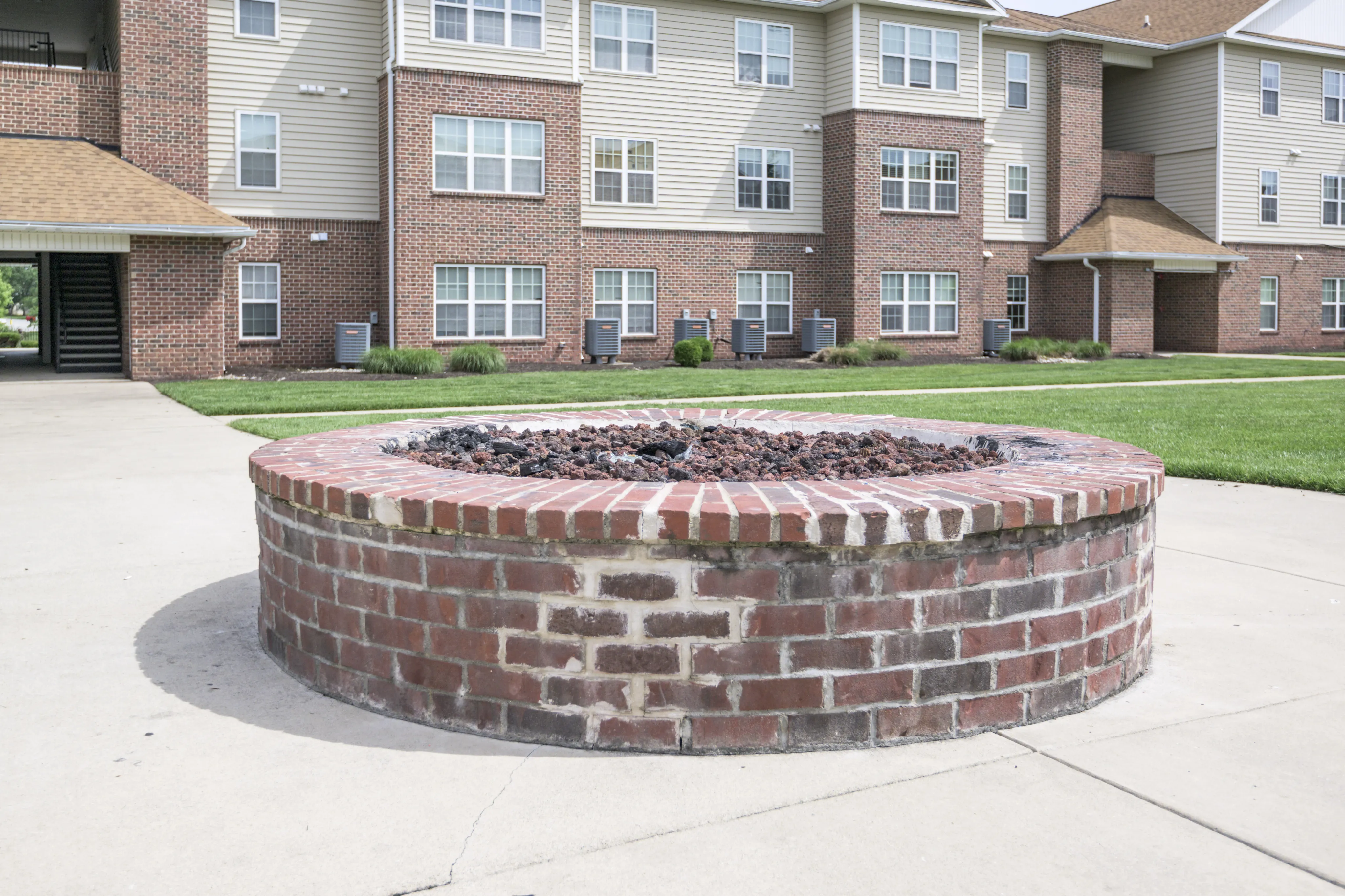 Charming brick apartment complex with a welcoming fire pit centerpiece and lush green surroundings.
