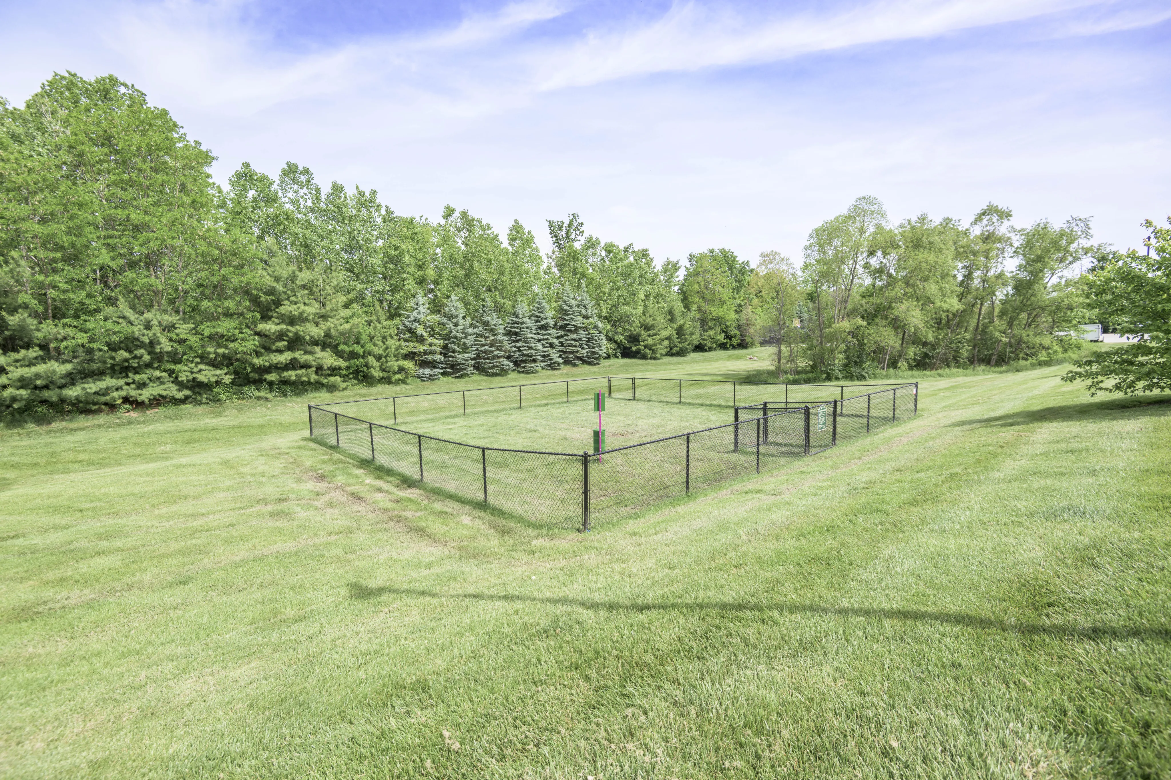 Expansive grassy area with a fenced-in section, surrounded by lush trees under a clear blue sky.